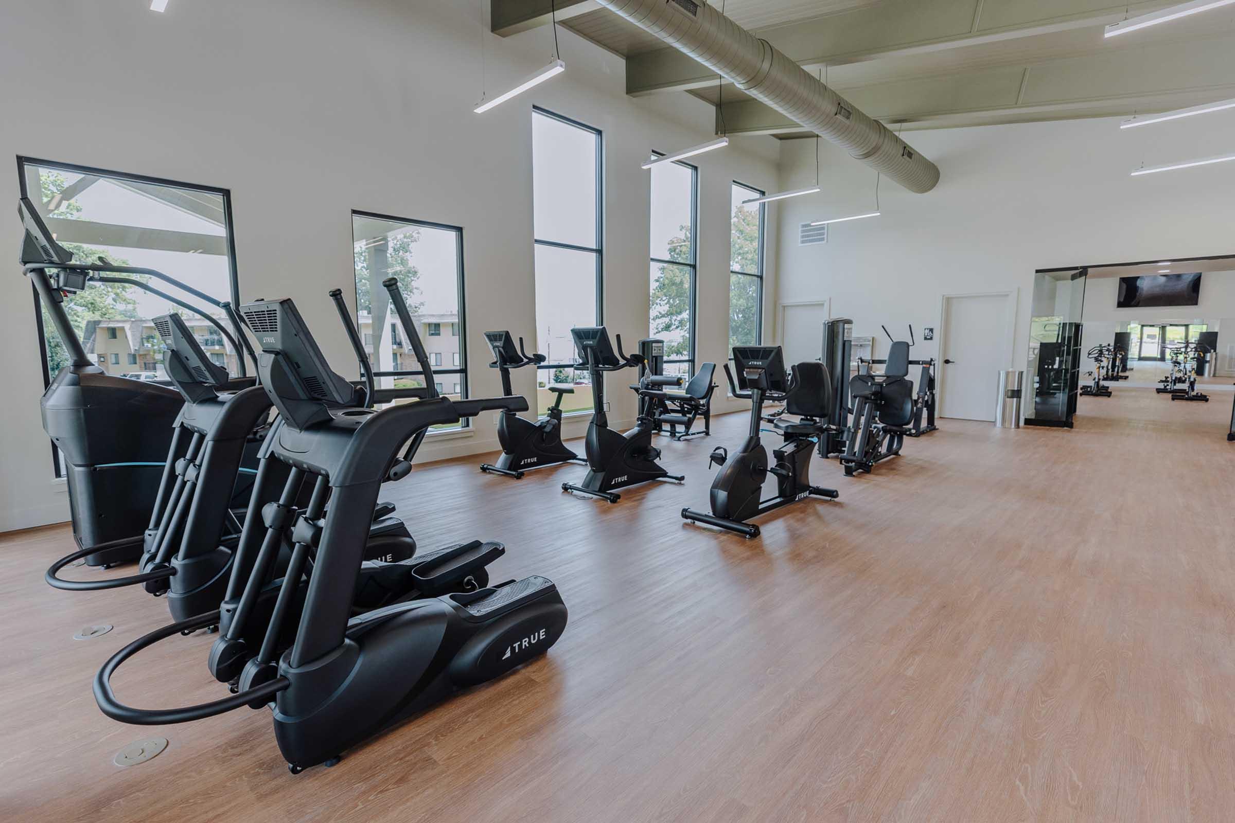A modern gym interior featuring several pieces of exercise equipment, including elliptical machines and stationary bikes. Large windows allow natural light to illuminate the space, and the flooring is wooden. The atmosphere is bright and inviting, designed for fitness activities.