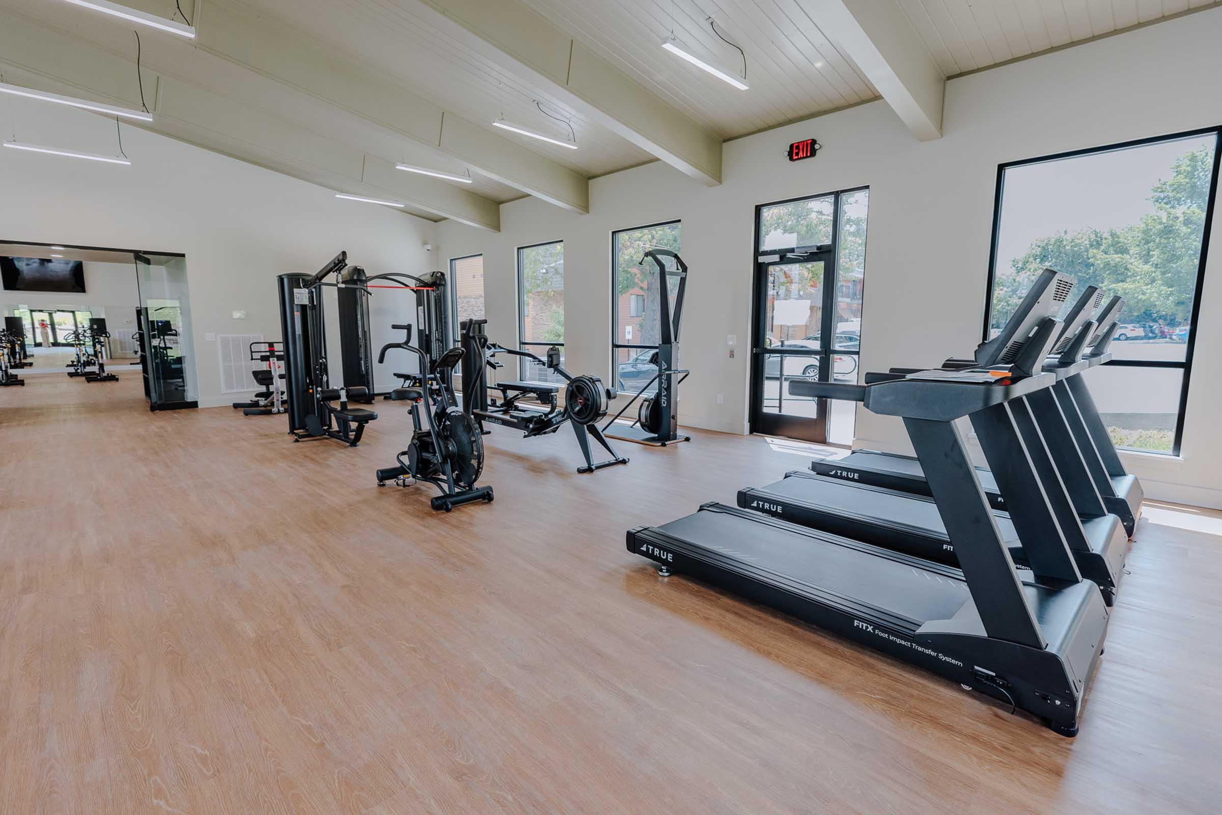 A modern gym interior featuring several pieces of fitness equipment, including treadmills and strength training machines, with large windows allowing natural light to fill the space. The wooden floor and bright walls create a welcoming atmosphere for workouts.