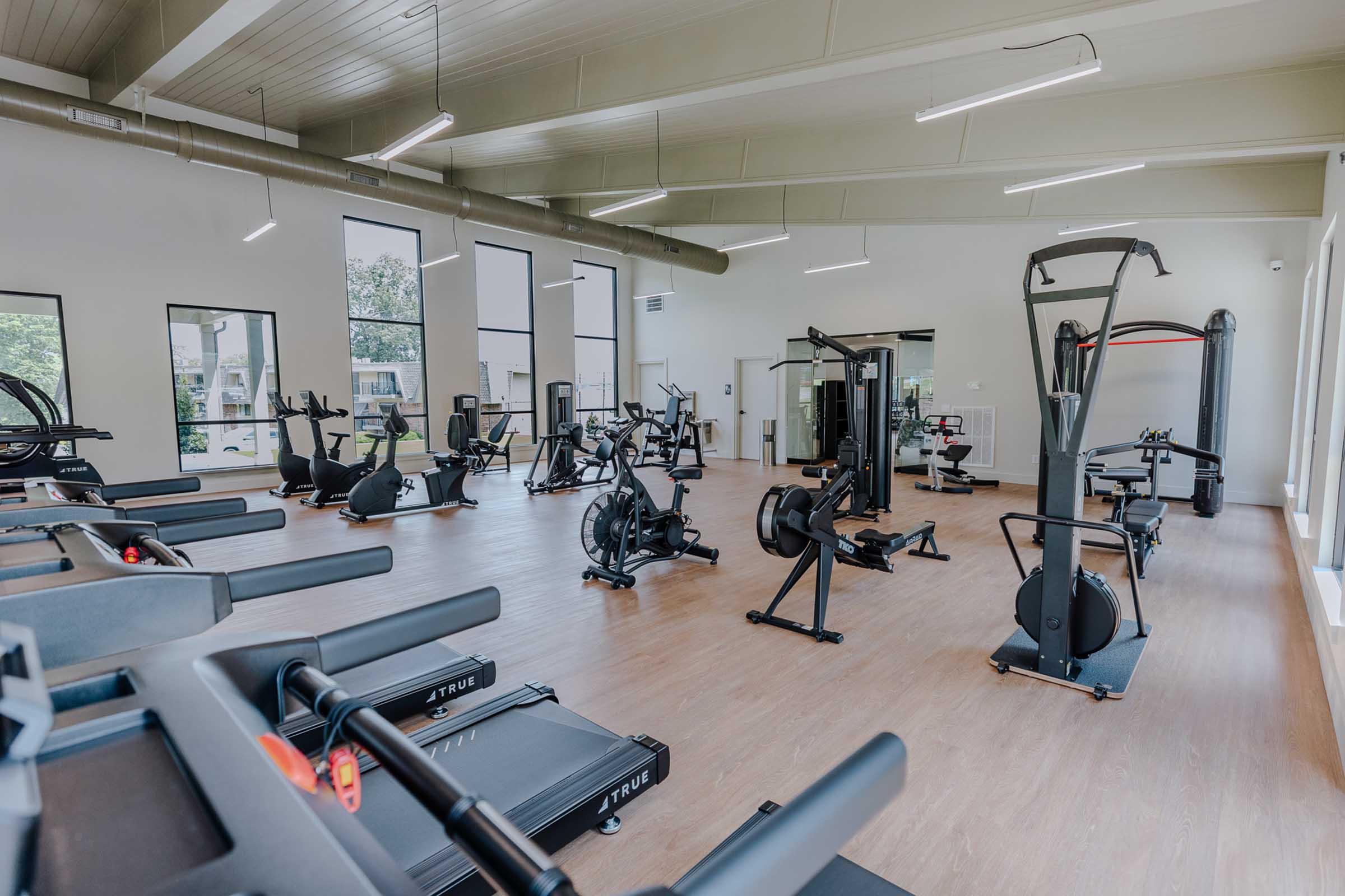 A spacious, modern gym featuring various exercise equipment, including treadmills, stationary bikes, and weight machines. Large windows provide natural light, and the flooring is hardwood. The gym appears well-maintained and inviting for fitness enthusiasts.