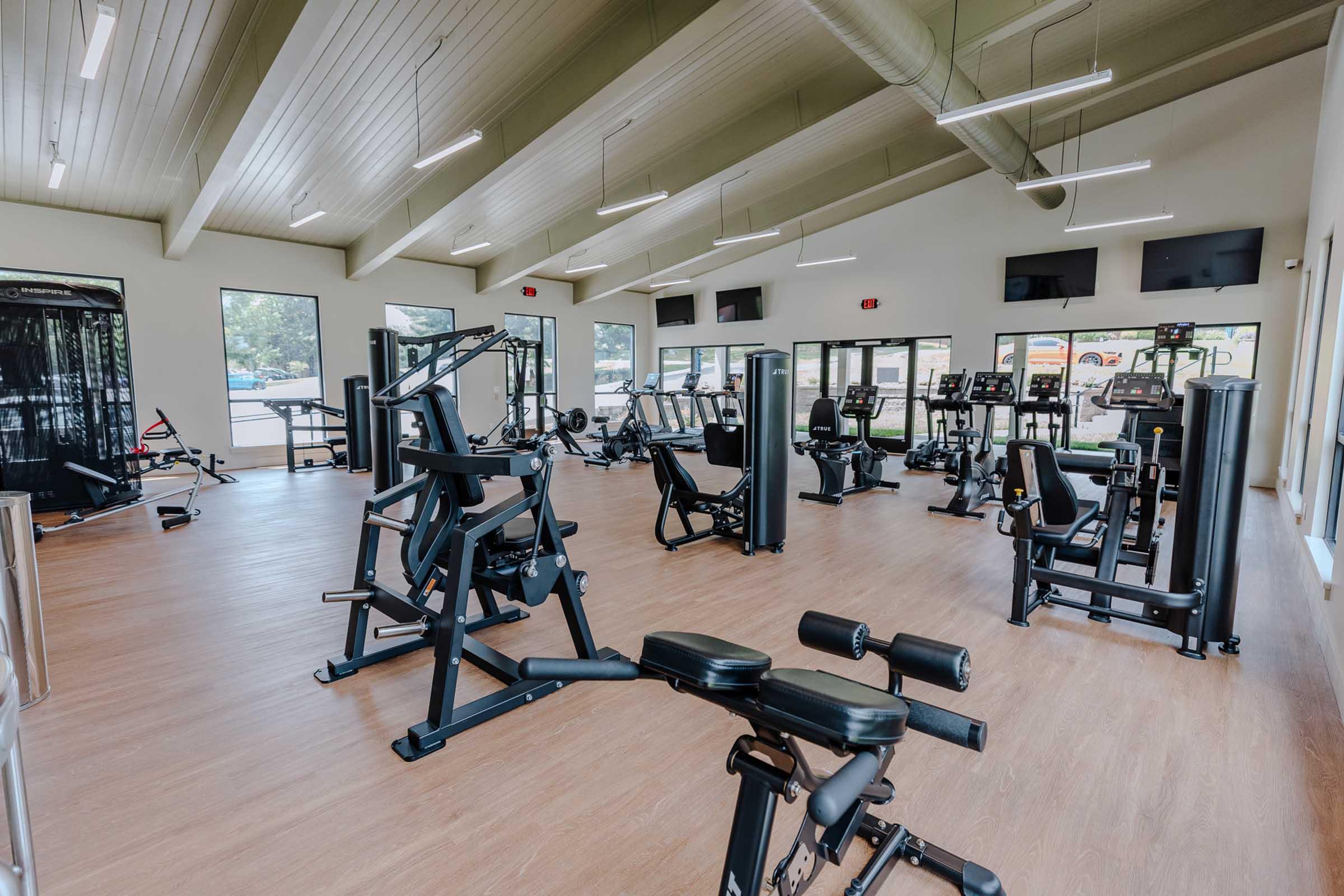 A modern gym interior featuring various fitness equipment including weight machines, cardio machines, and benches. The space is well-lit with large windows and a high ceiling, creating an inviting atmosphere for workouts. There are multiple display screens mounted on the walls.