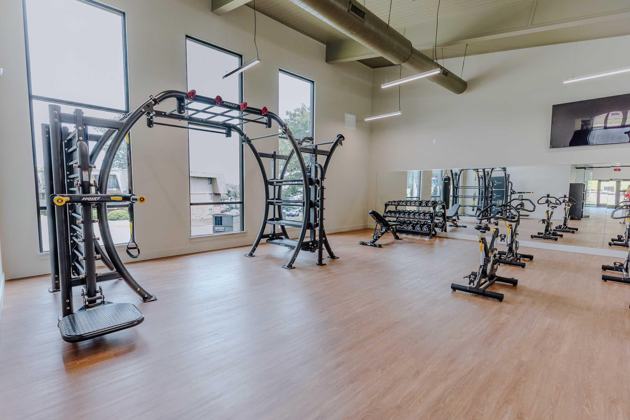 A spacious gym interior featuring exercise equipment including weight machines and dumbbells. Large windows allow natural light to fill the room, while a mirrored wall enhances the sense of space. A cable machine is prominently displayed in the center, with a wooden floor adding a warm touch to the environment.