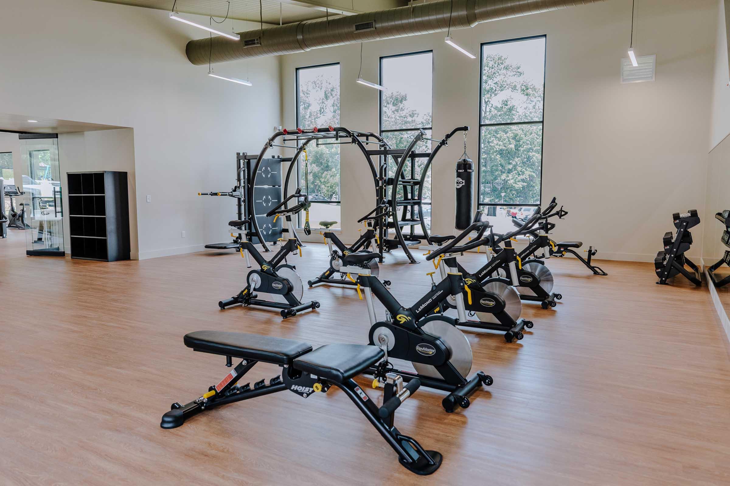 A modern fitness studio featuring various exercise equipment. In the foreground, there are benches and stationary bikes. The background includes a multi-gym setup, a punching bag, and large windows allowing natural light. The floor is wooden, creating a warm and inviting atmosphere for workouts.