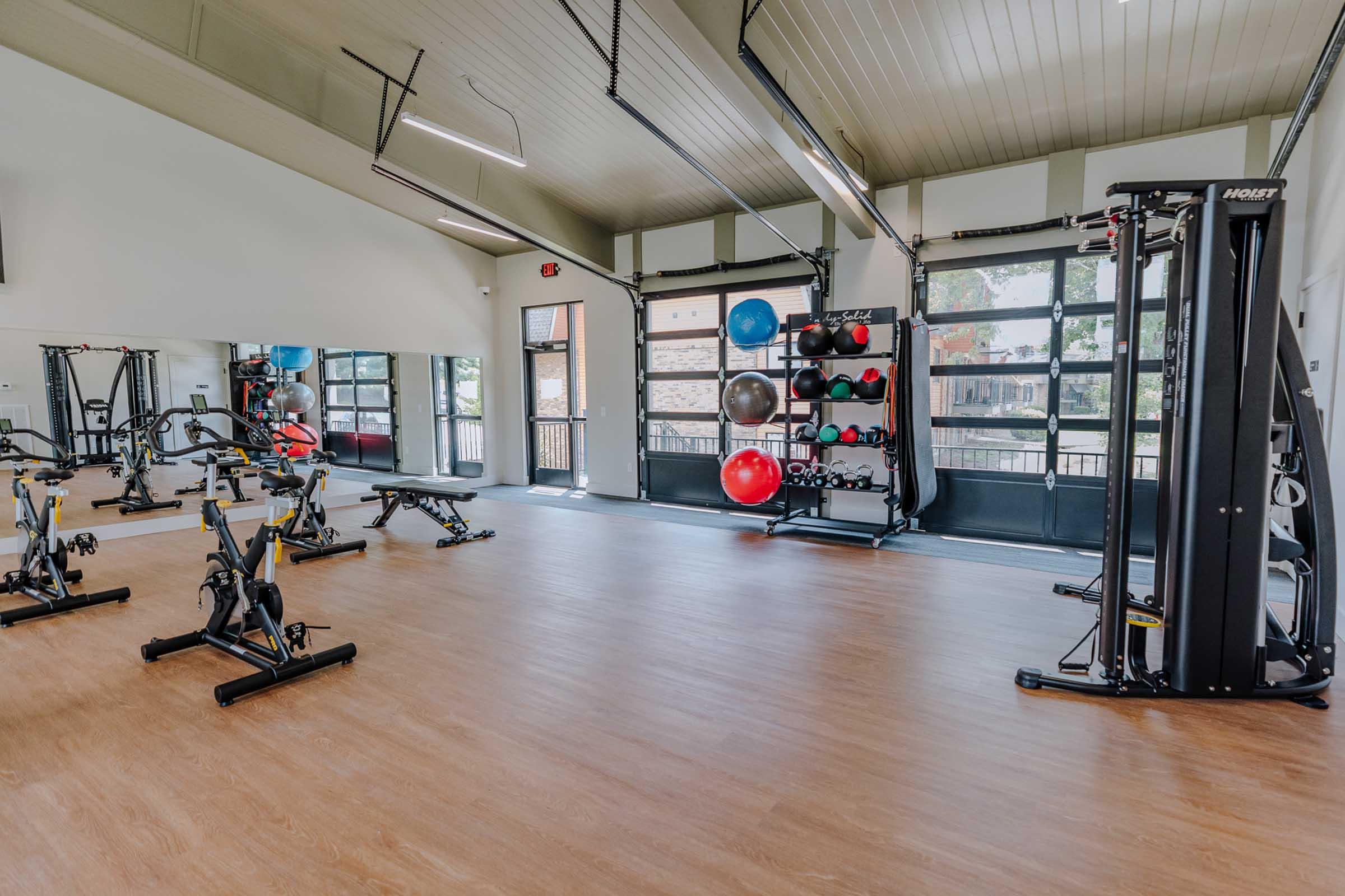 A modern gym interior featuring various exercise equipment, including weight machines, benches, and stability balls. The space has large windows allowing natural light, wooden flooring, and a clean, organized layout, creating an inviting atmosphere for workouts.