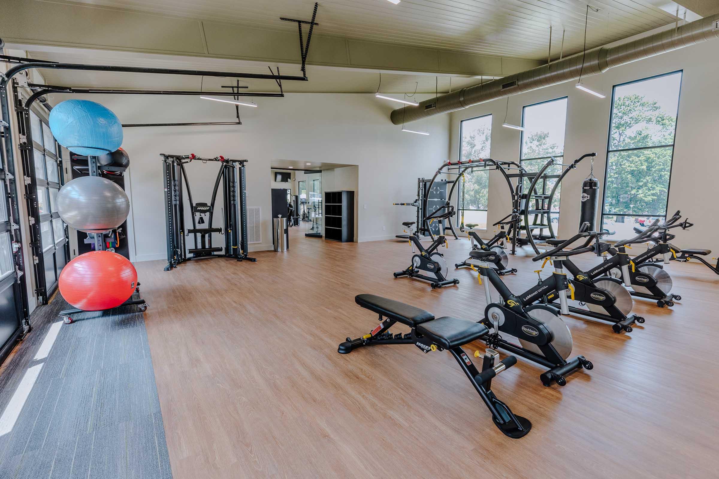 A spacious gym interior featuring various exercise equipment, including weight machines, stationary bikes, and free weights. Bright windows allow natural light to fill the room, enhancing the modern and inviting atmosphere. Fitness balls are neatly arranged in the corner, contributing to a well-equipped workout space.