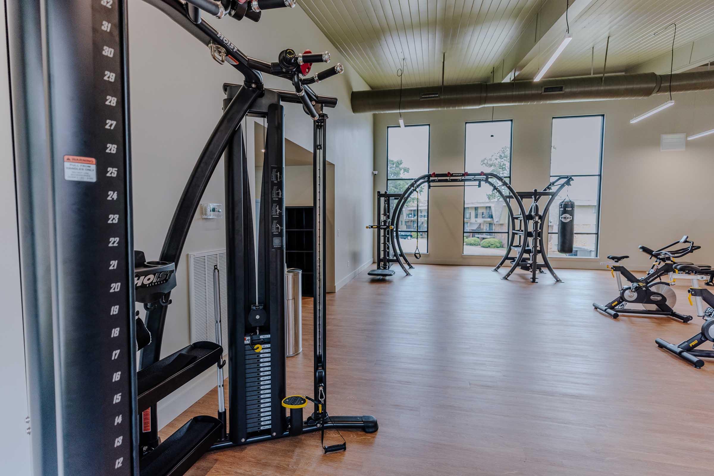 Interior view of a modern gym featuring various exercise equipment, including cable machines and stationary bikes. Large windows provide natural light, illuminating the spacious area with wooden flooring. The gym is tidy and ready for workouts.