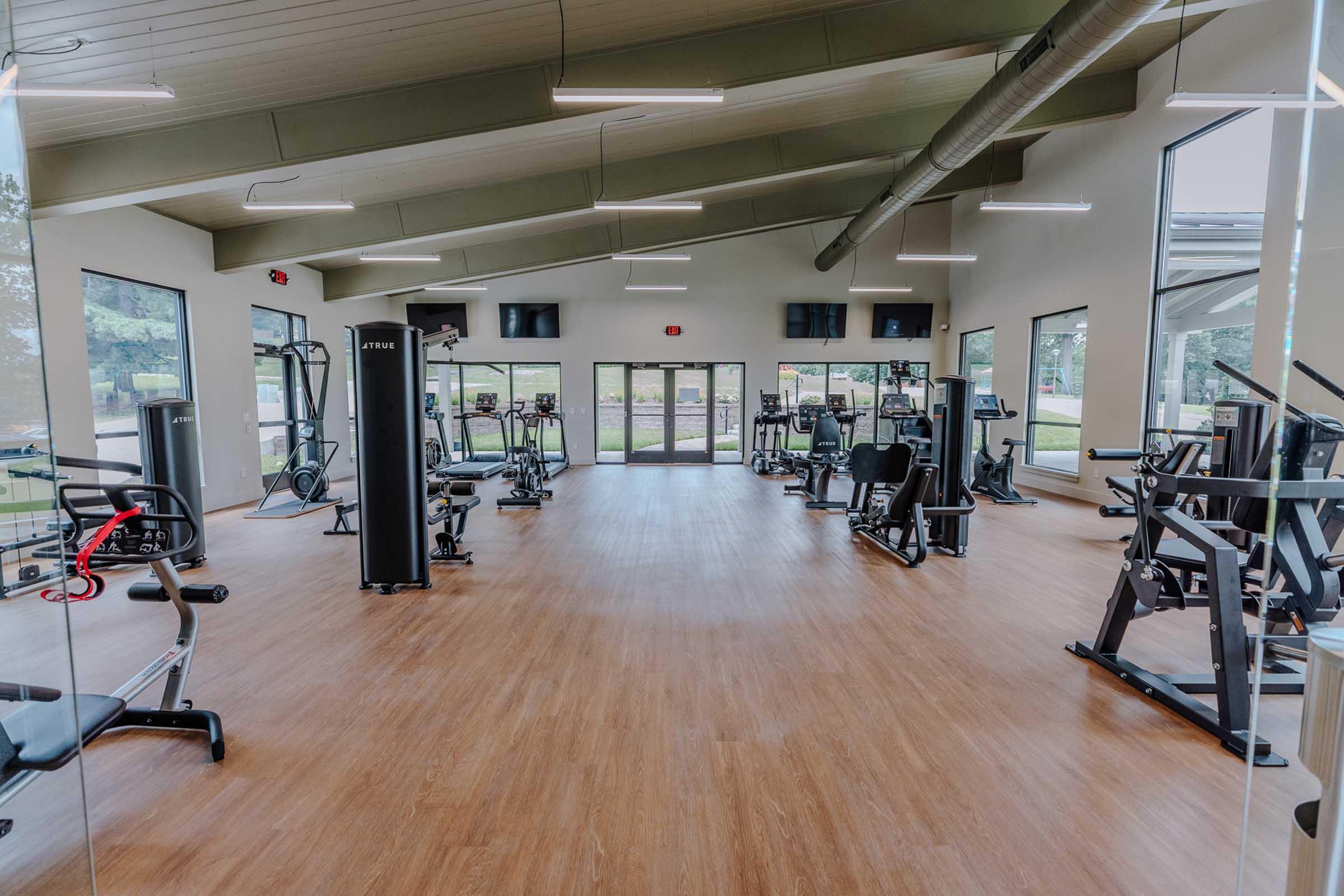 A spacious gym interior featuring various exercise equipment, including weight machines and cardio machines, set in a well-lit environment with large windows. The flooring is wooden, and there are multiple screens on the walls, creating a modern fitness space.