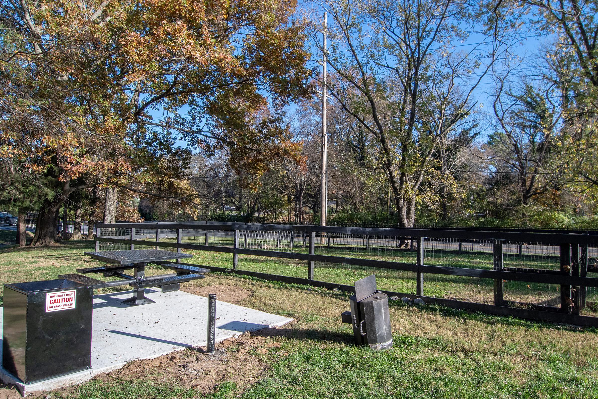 picnic area featuring a concrete pad with a picnic table, surrounded by grass and trees displaying autumn leaves. A caution sign is visible on a nearby structure, and a grill is placed next to the picnic table, all set against a clear blue sky.