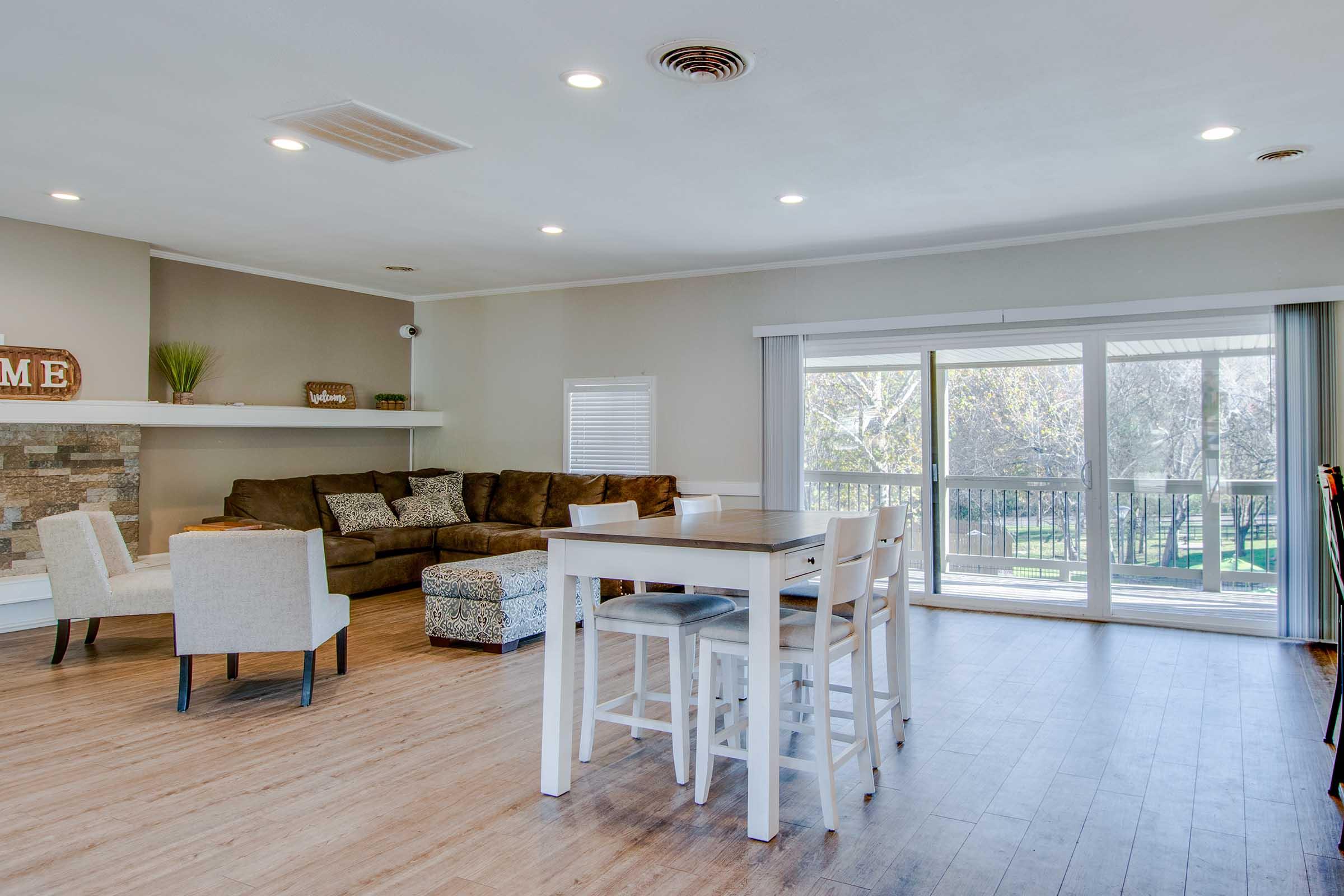 A cozy living area featuring a brown sectional sofa and light-colored chairs. There is a wooden dining table with white chairs and a stone accent wall. Large sliding glass doors open to a view of greenery outside, allowing natural light to fill the space. The décor is simple and inviting.