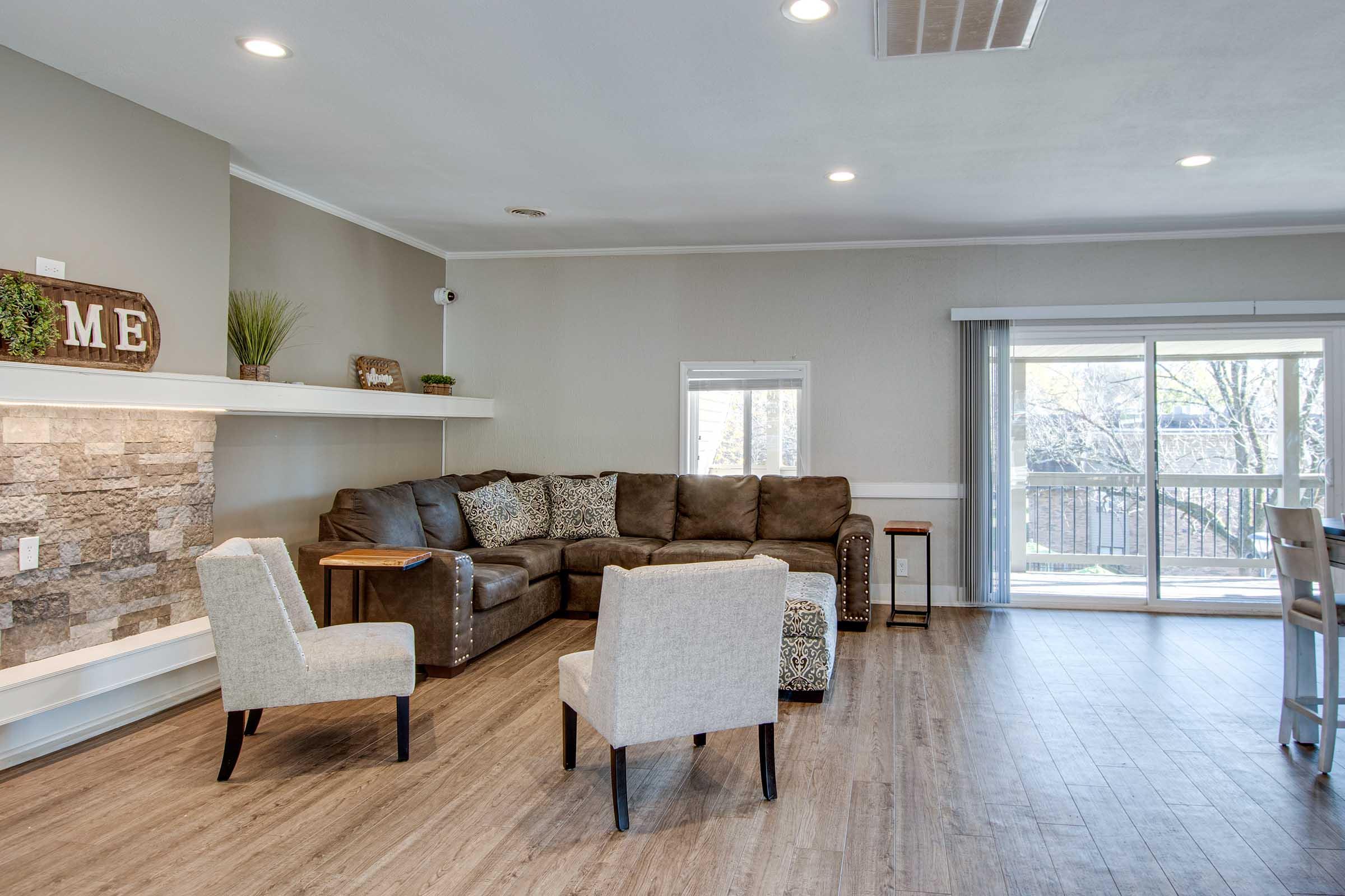 A cozy living room featuring a brown sectional sofa with decorative pillows, two light-colored armchairs, a small side table, and a stone fireplace. Large windows provide natural light and a view of outdoor greenery. A wooden shelf displays decorative items, and the room has a modern, inviting atmosphere.