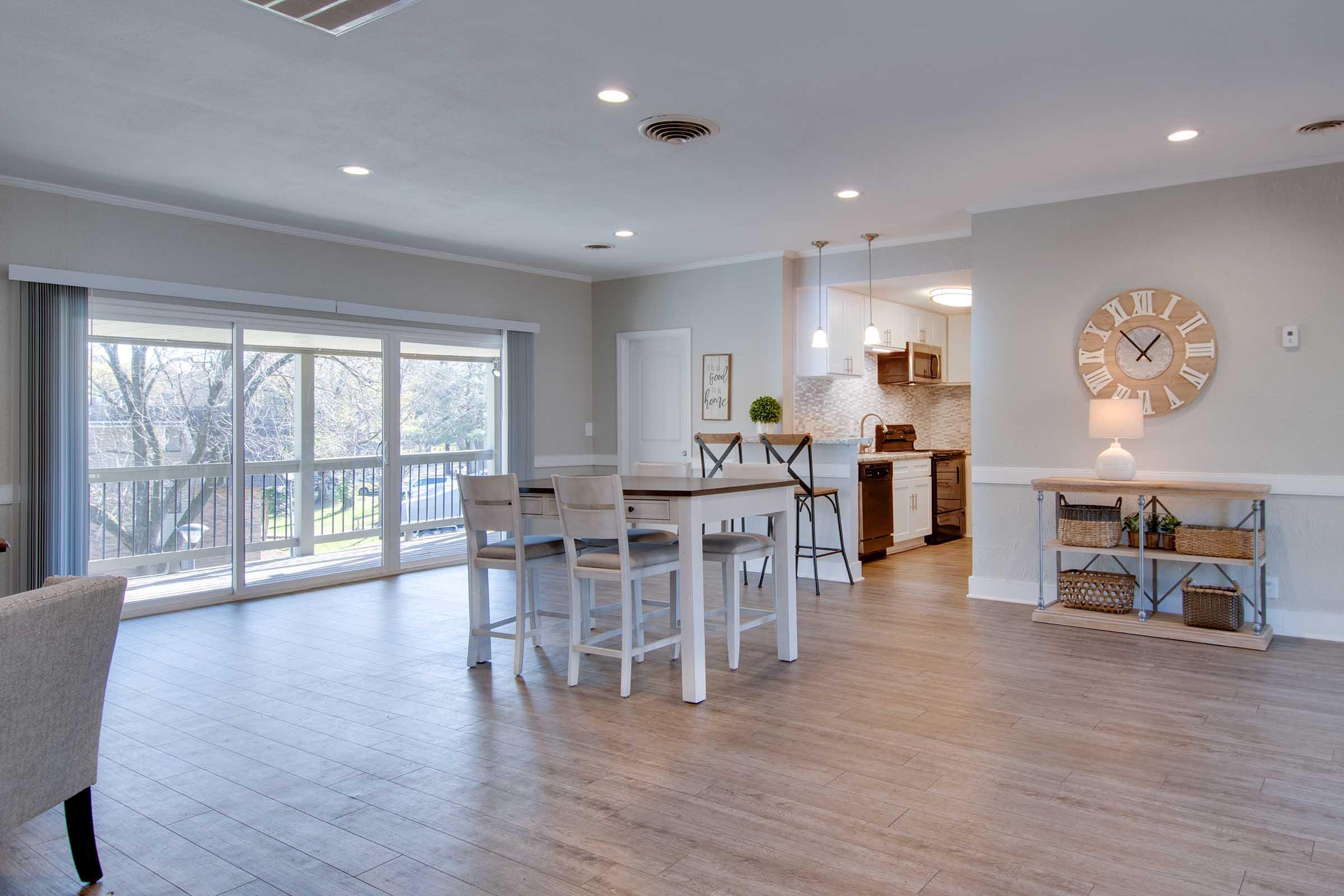 A bright and modern living space with a dining area featuring a white table and chairs. The kitchen is visible in the background, showcasing wood cabinetry and a stylish backsplash. Large windows let in natural light, offering a view of trees outside. A decorative clock and storage baskets add charm to the room.