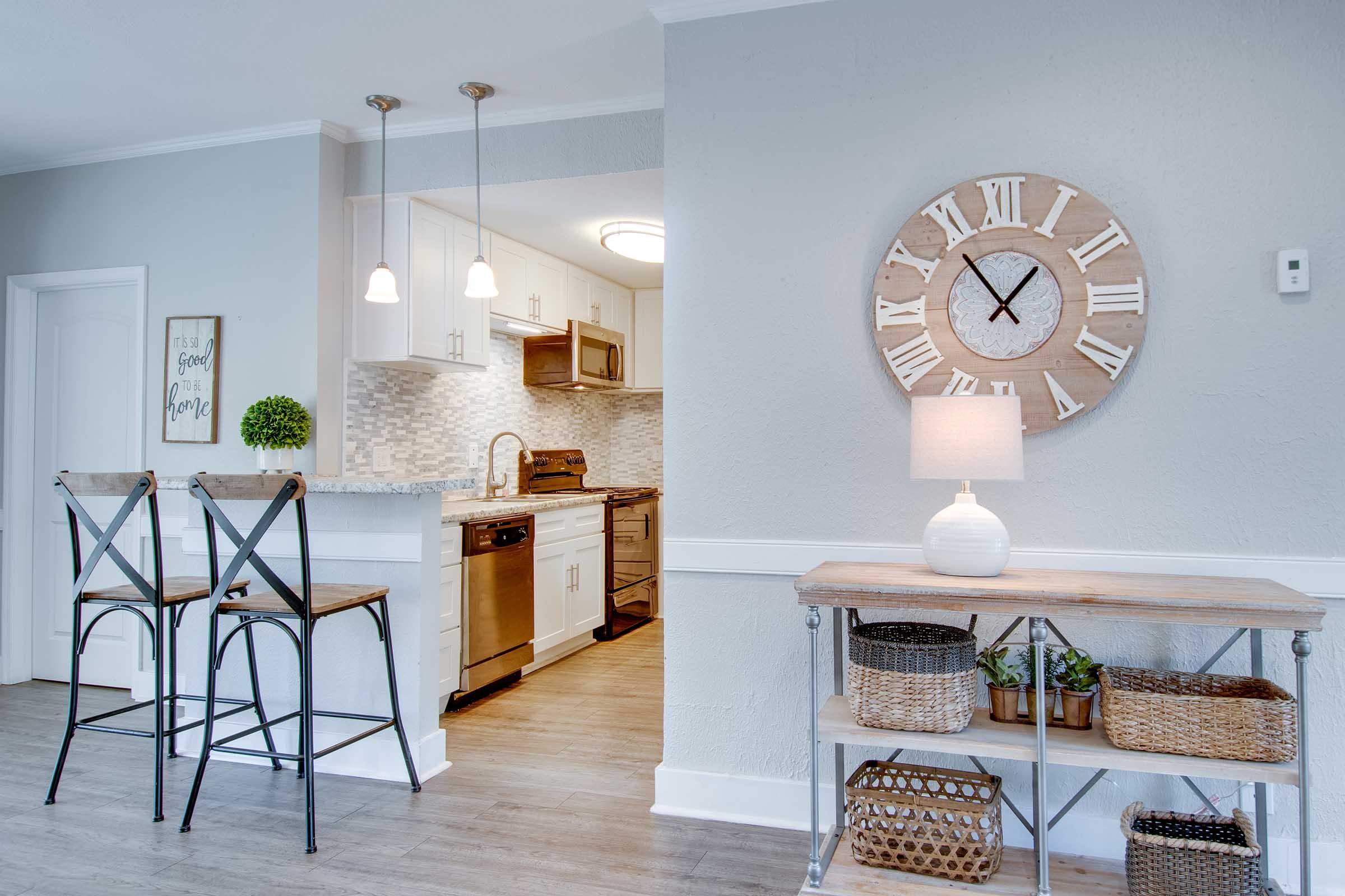 A modern kitchen and dining area featuring a gray wall, wooden accents, and stylish decor. The kitchen includes stainless steel appliances, a light backsplash, and a breakfast bar with two stools. A large wall clock and a lamp sit on a table with woven baskets, enhancing the inviting atmosphere of the space.