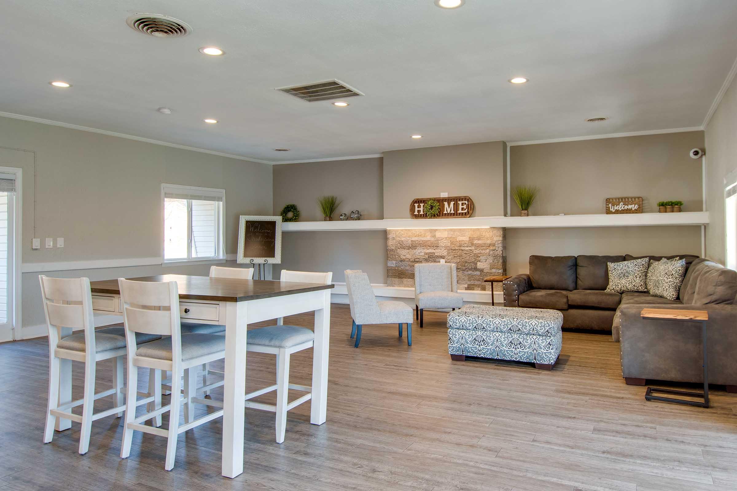A modern living space featuring a wooden dining table with white chairs, a cozy seating area with a brown couch, decorative pillows, and an ottoman. The walls are a soft gray, and there’s a stone accent wall with a “HOME” sign. Natural light floods in from large windows, enhancing the warm ambiance.