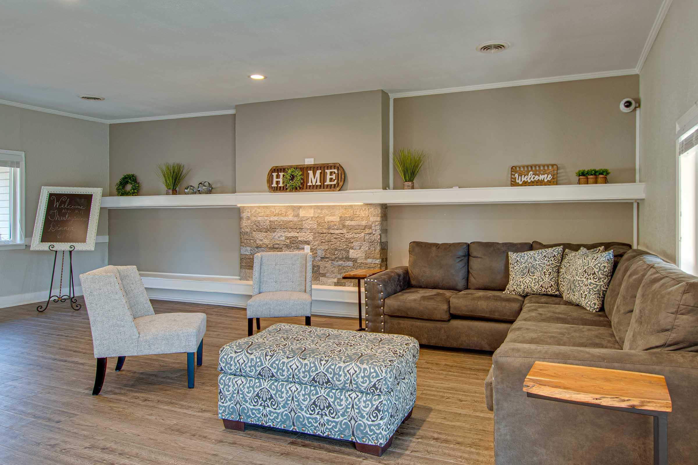 A cozy living room featuring a large sectional sofa with decorative pillows, two upholstered chairs, and an ottoman. The space is enhanced by a stone fireplace with a wooden mantle displaying decorative accents. Soft lighting and potted plants add warmth to the inviting atmosphere. A welcome sign is also visible.