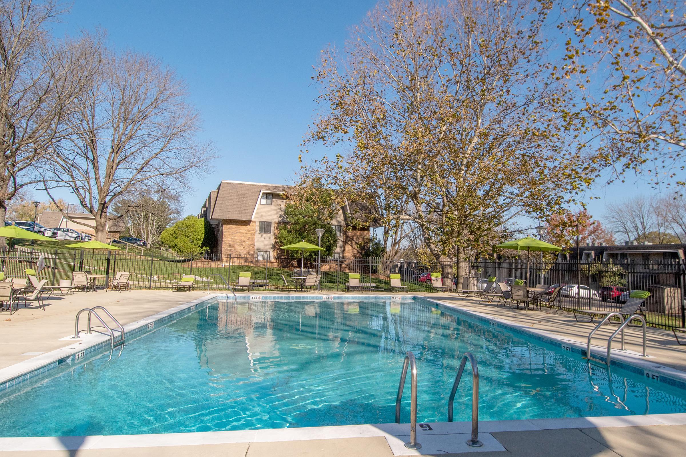 A clear blue swimming pool surrounded by lounge chairs and green umbrellas. In the background, there are apartment buildings and trees with some fall foliage. The sky is bright and clear, indicating a sunny day.