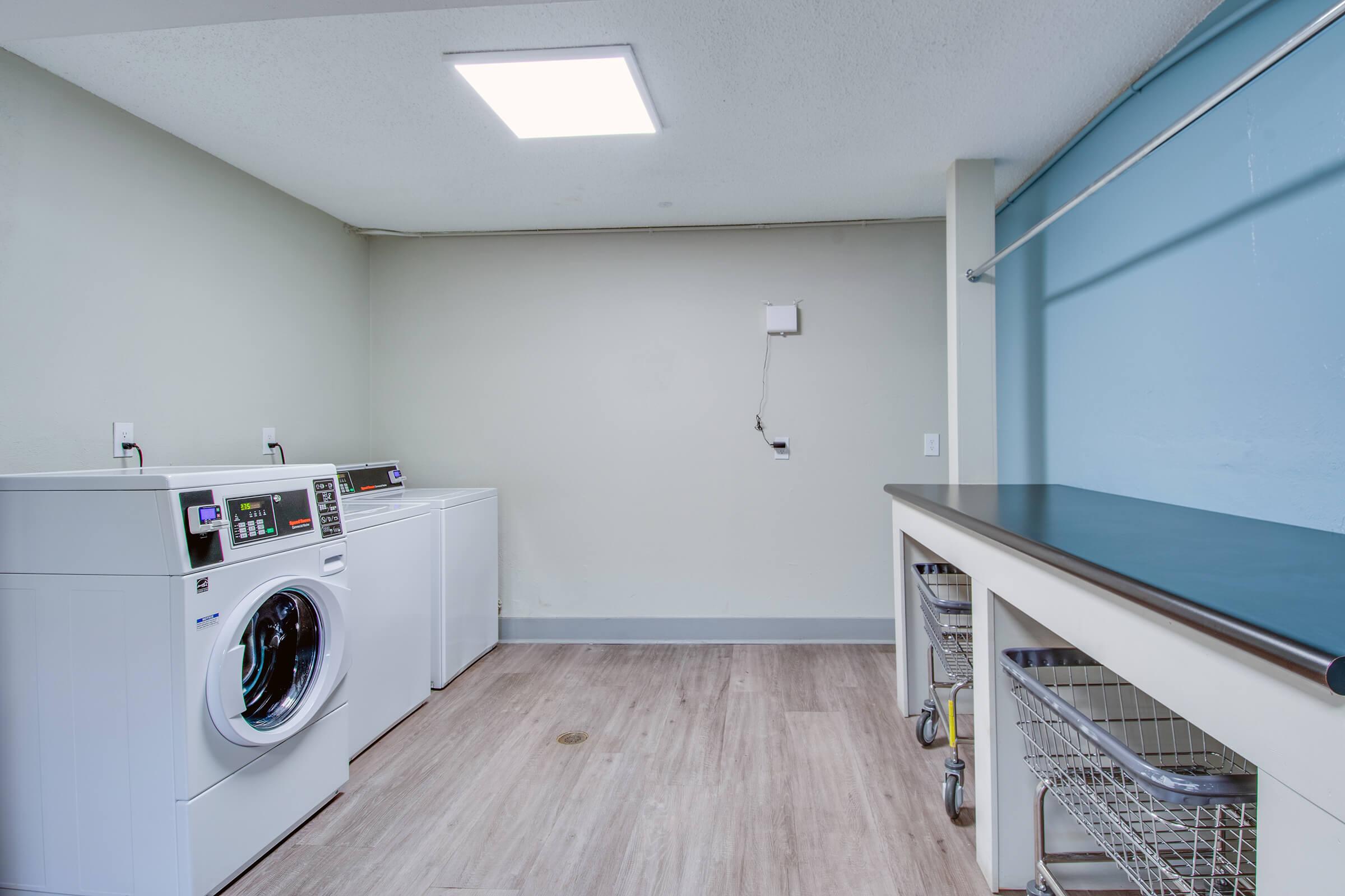Modern laundry room featuring two washing machines, a towel drying rack, and a countertop with storage carts. The walls are painted in light colors, and overhead lighting illuminates the space, creating a clean and organized environment.