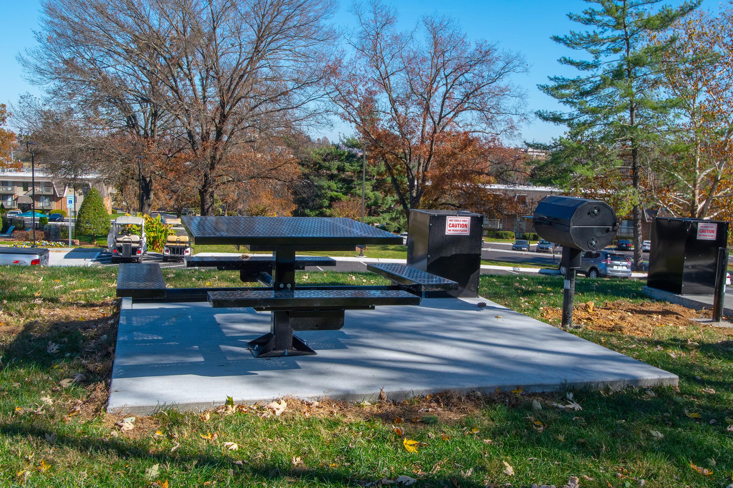 A picnic area featuring a black, metal picnic table and grill set on a grassy patch with fallen leaves. Surrounding trees show autumn colors, and a residential area is visible in the background. Caution signs are affixed to the picnic area structure.