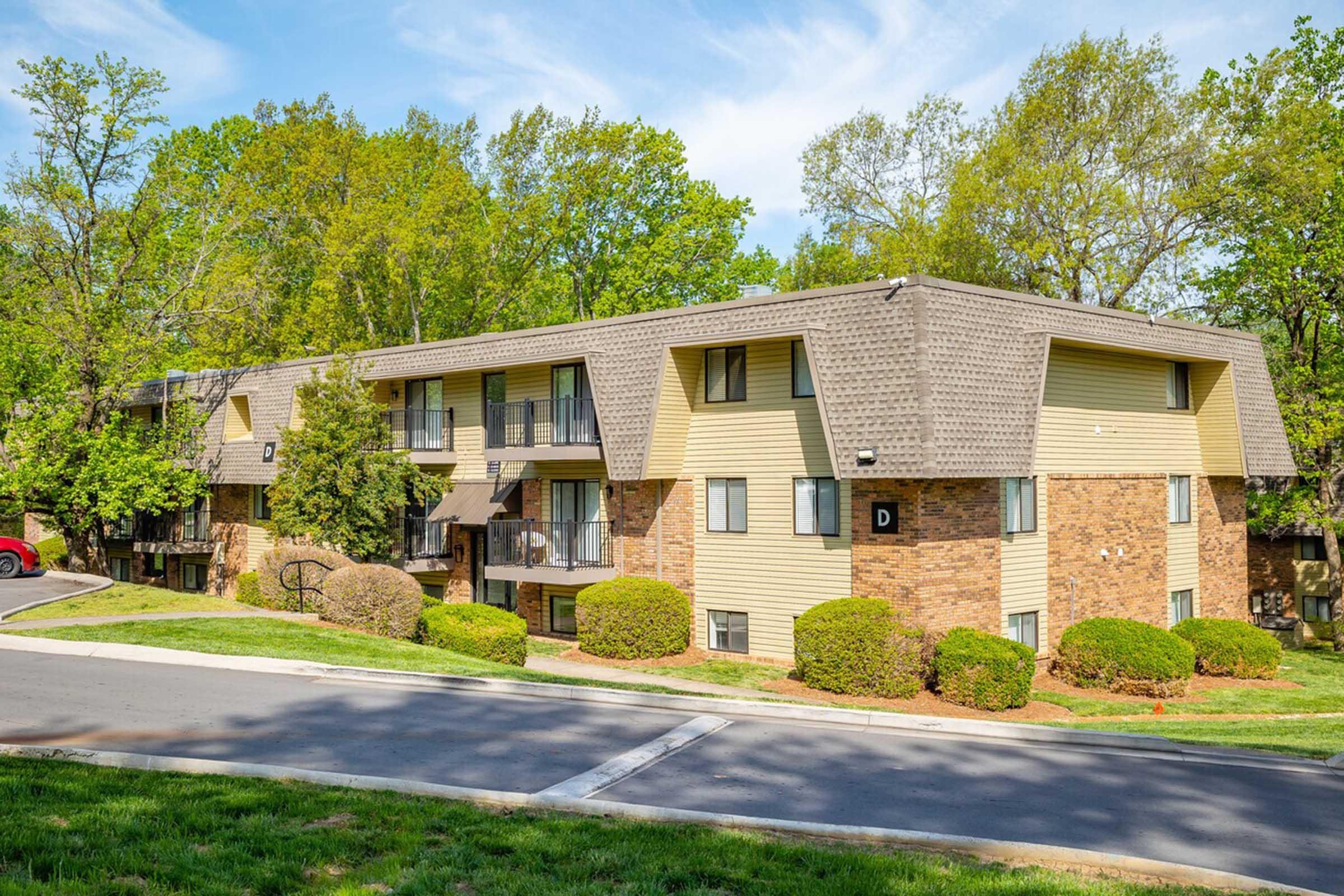 Three-story apartment building with a mix of brick and siding exterior, featuring landscaped bushes and trees in the background. The building has balconies and is situated alongside a paved road and grassy area. Clear blue sky visible above.