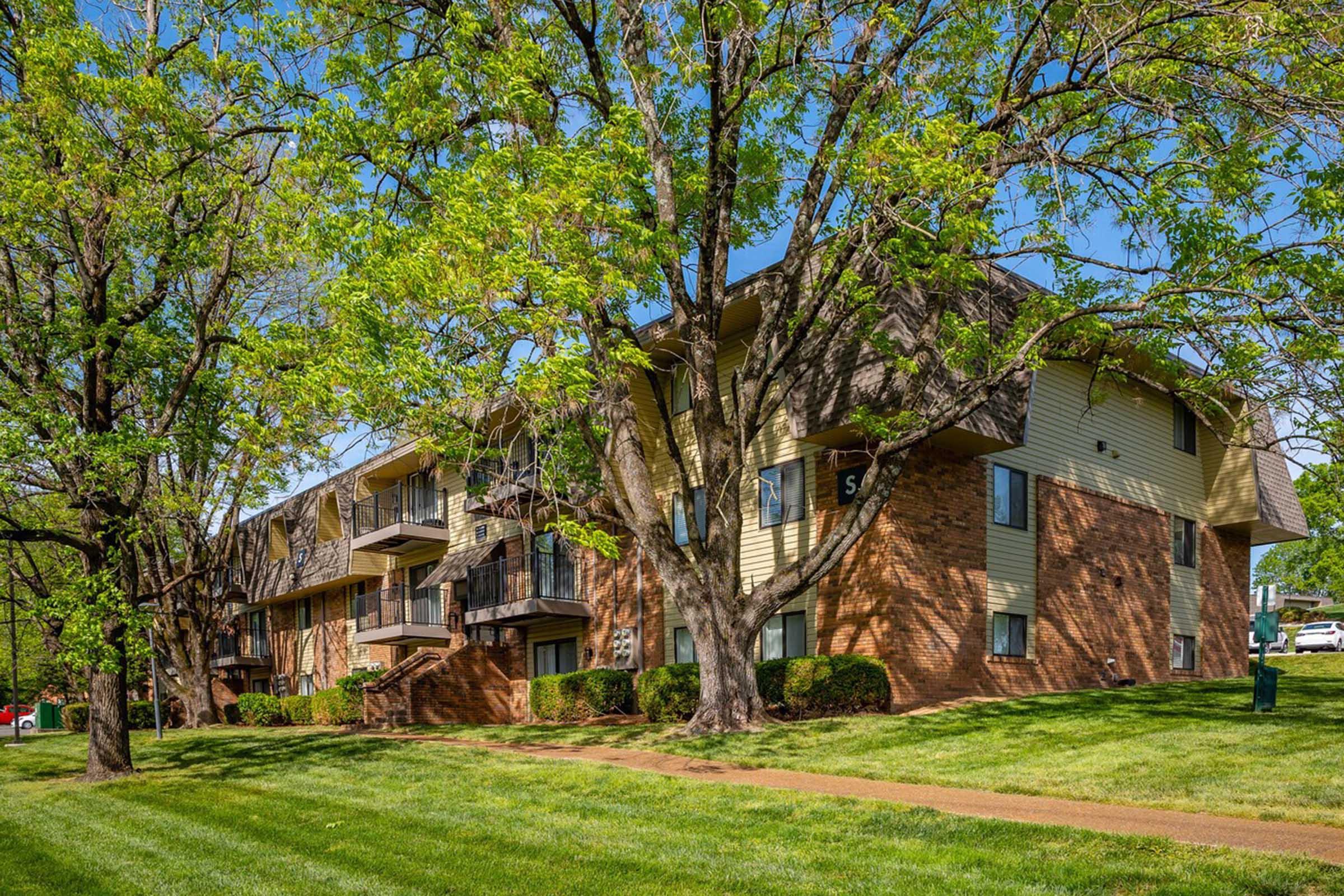 A multi-story apartment building with brick and wooden siding, surrounded by lush green lawns and trees. The scene features a clear blue sky and well-maintained landscaping, showcasing a peaceful residential area. Balconies are visible on the upper floors, adding to the inviting atmosphere.