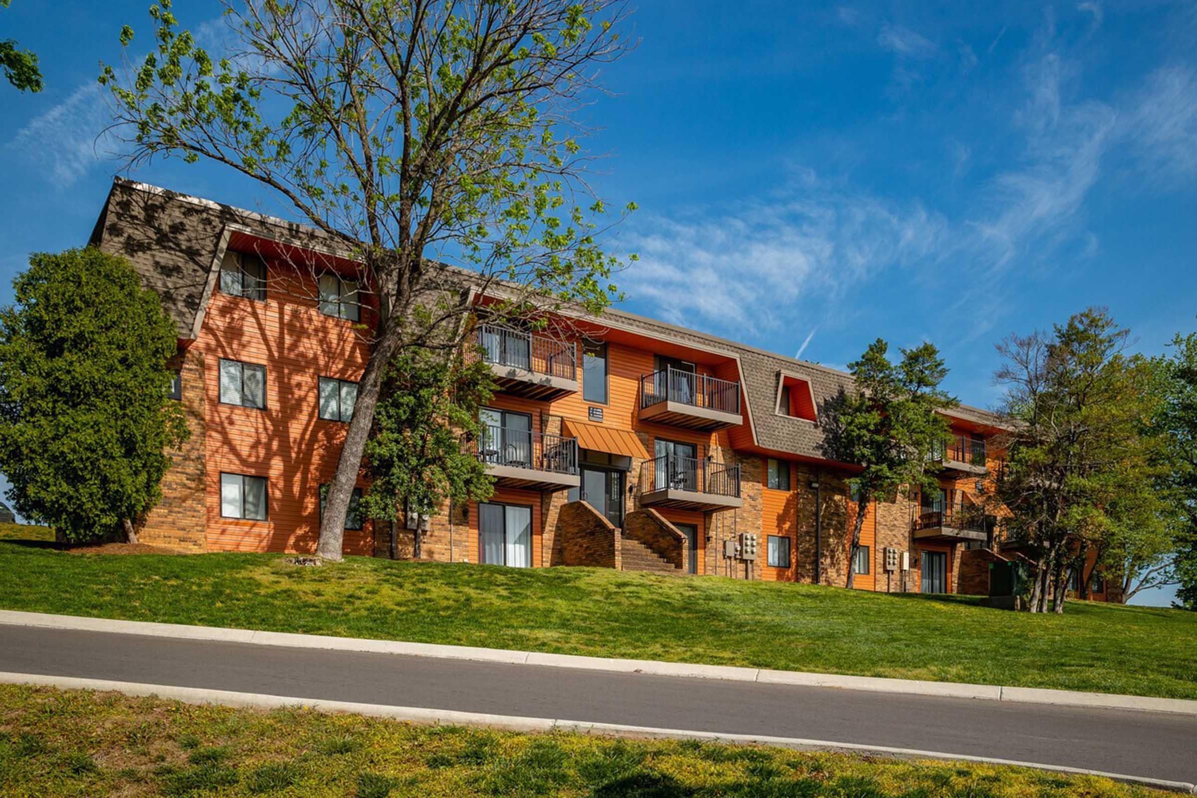 A multi-story building with a mix of stone and orange siding, featuring balconies and a grassy area in front. Several trees surround the building under a clear blue sky. The structure is set back from the road, which is lined with a neat curb.
