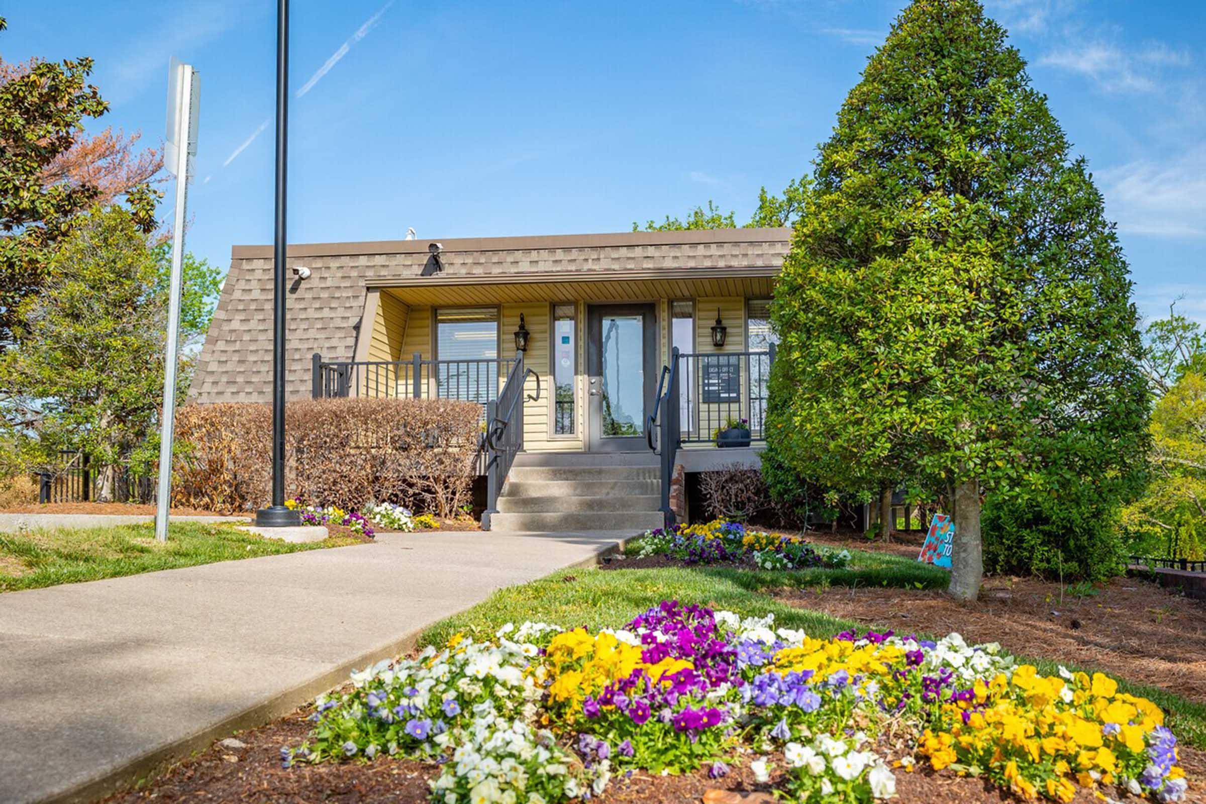 A charming building with a sloped roof, featuring a welcoming front entrance and surrounded by vibrant flower beds filled with yellow, purple, and white blooms. Lush green trees and shrubs are in the background under a clear blue sky. A lamppost stands to the left of the path leading to the entrance.