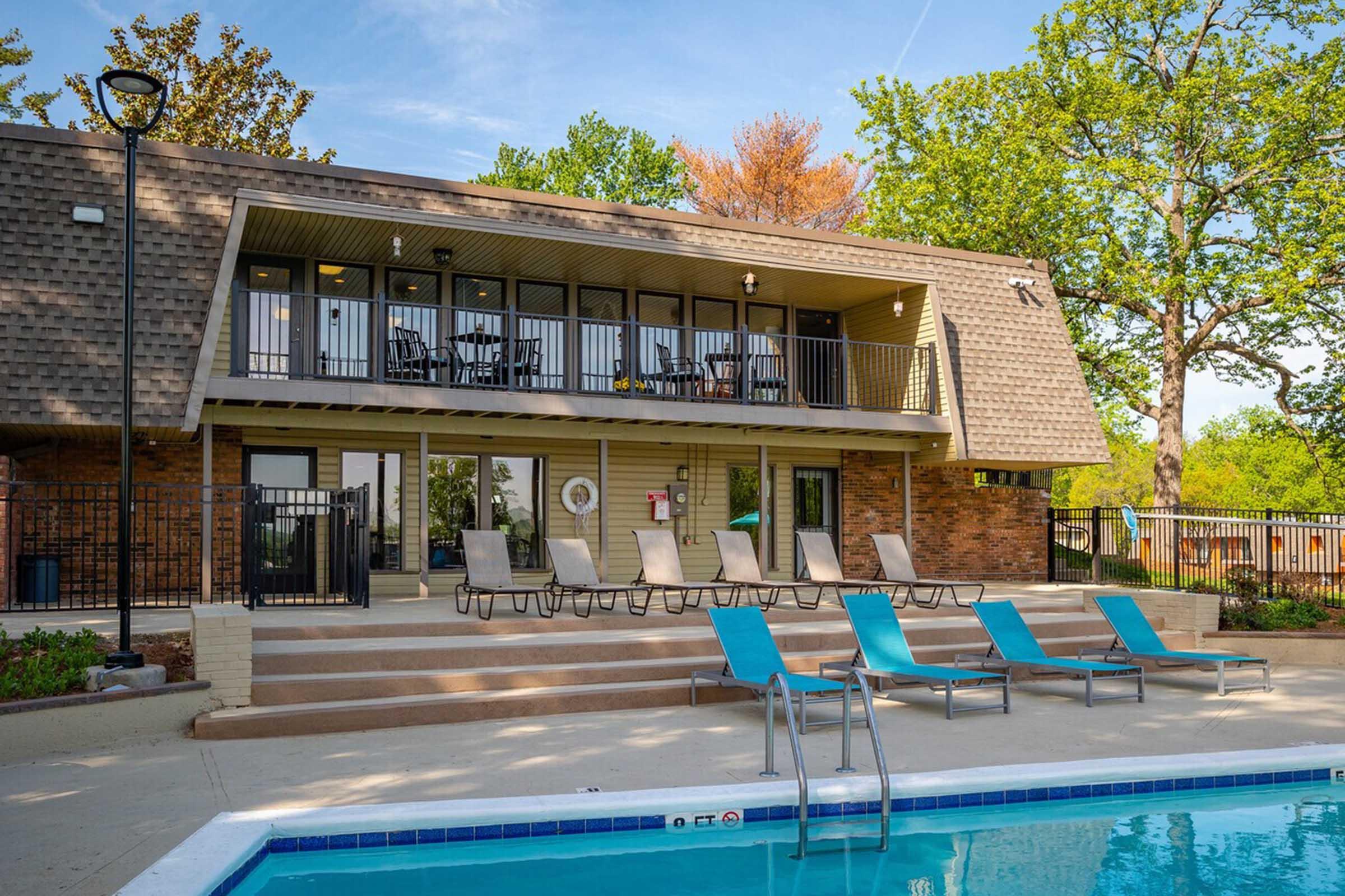 A view of a pool area with several lounge chairs lined up beside the water, and a building with a balcony overlooking the pool. The setting is surrounded by trees and greenery, creating a relaxing outdoor atmosphere on a sunny day.