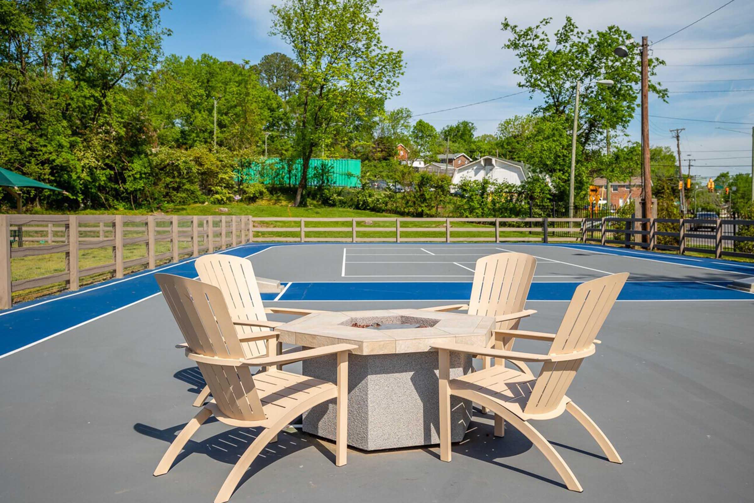 A view of a tennis court featuring a fire pit surrounded by four wooden chairs, with greenery and residential buildings in the background. The court is marked with blue and white lines under a clear blue sky, and wooden fencing outlines the area.