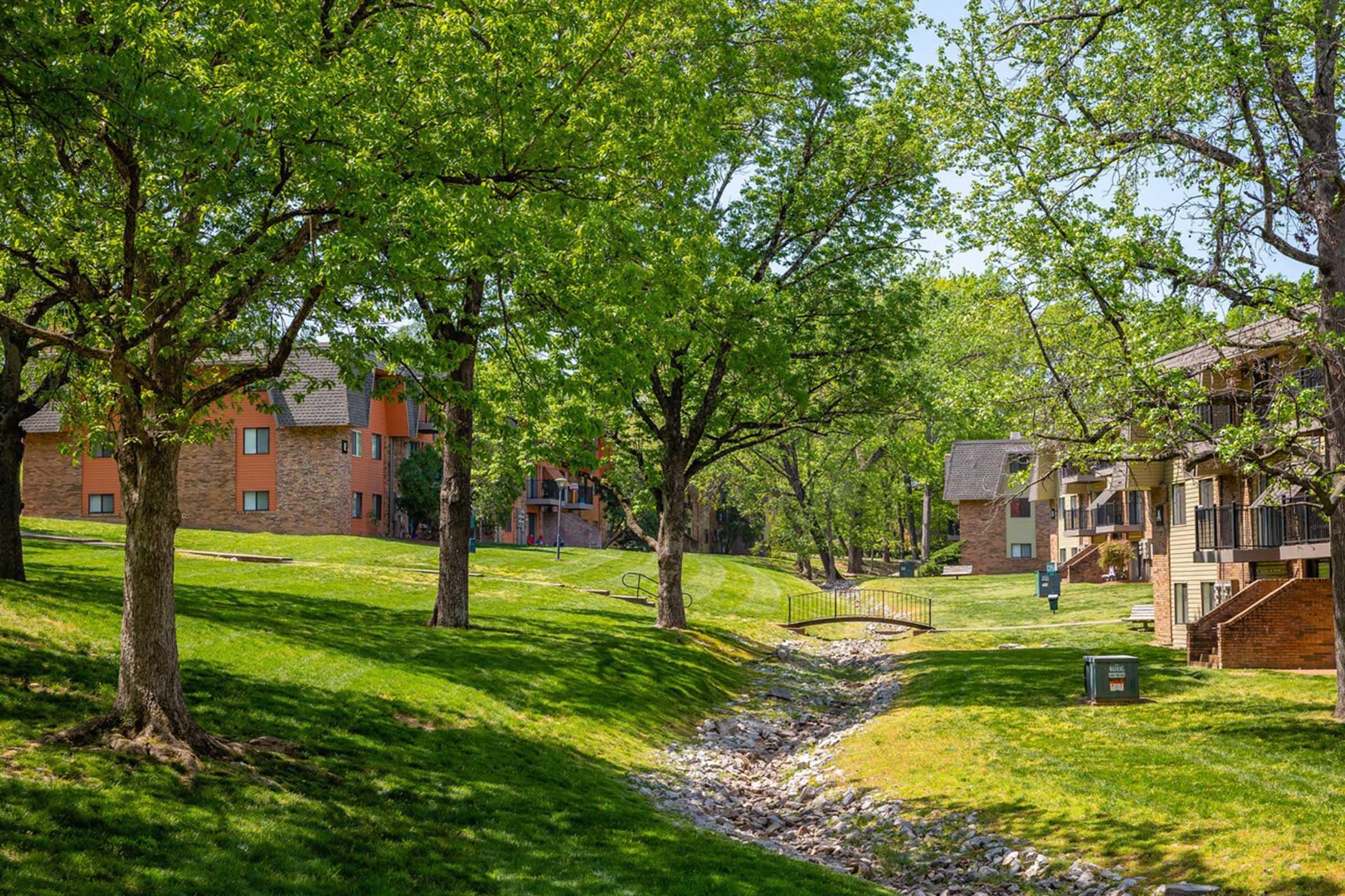 Lush green lawn with trees lining a small creek, alongside brick apartment buildings. The sun is shining, creating a vibrant outdoor scene. The setting suggests a peaceful residential area with well-maintained landscaping.