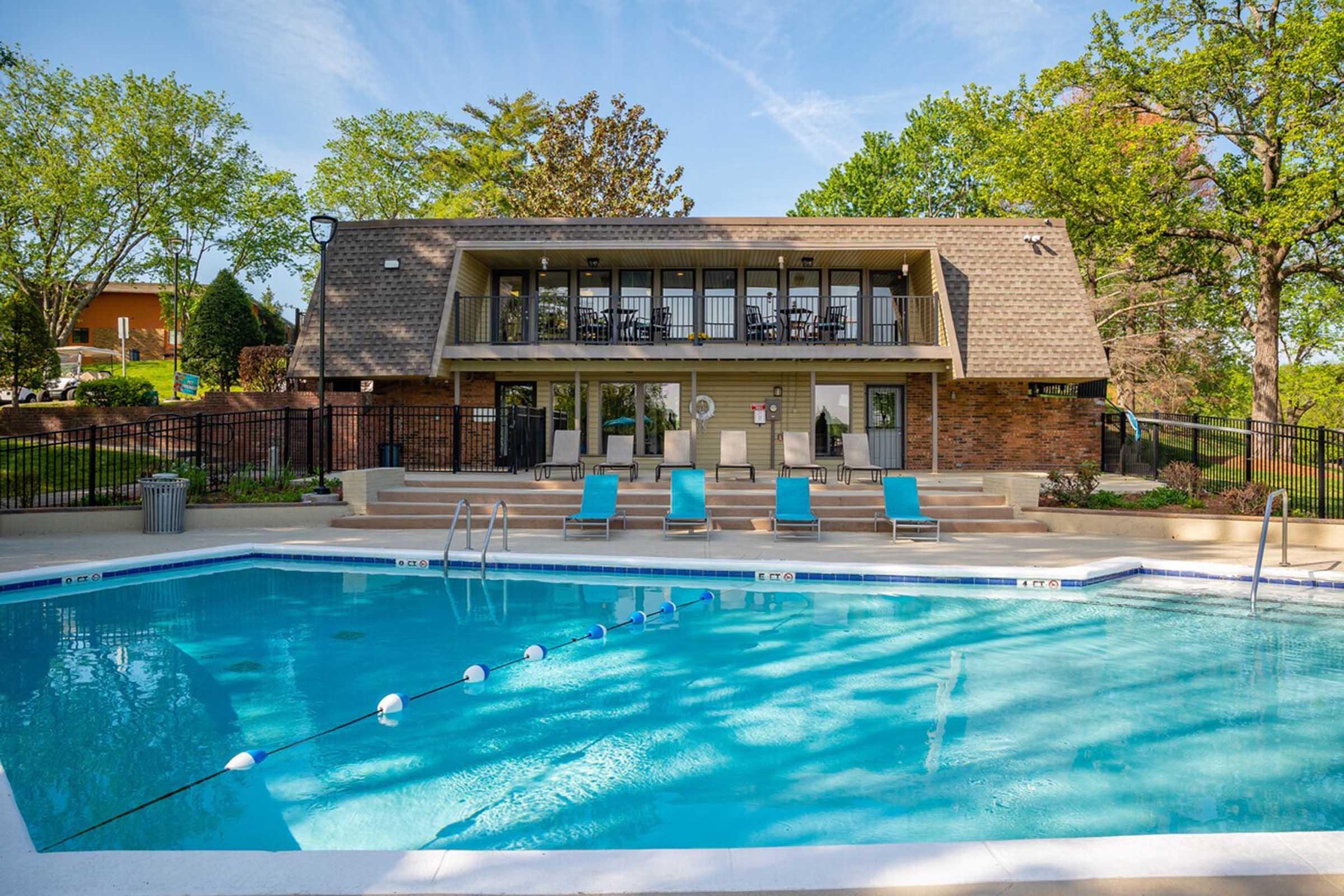 A clear blue swimming pool surrounded by lounge chairs, with a building featuring large windows in the background. The area is lush with green trees and plants, creating a serene environment. A safety float is visible in the pool, and the sky is bright with a few clouds.
