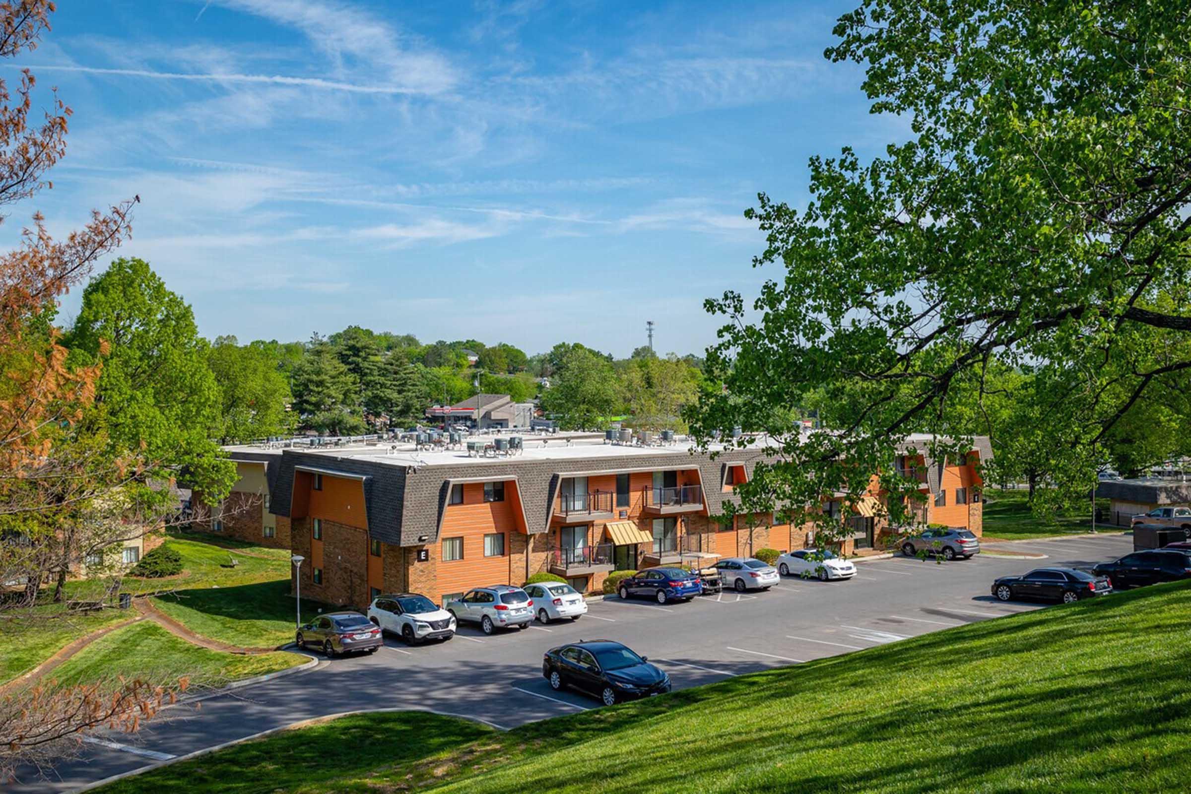 A view of a multi-story building with a landscaped yard, surrounded by trees and grass. Several cars are parked in front of the building, and the sky is clear with a few clouds. The setting appears to be a quiet residential or commercial area.