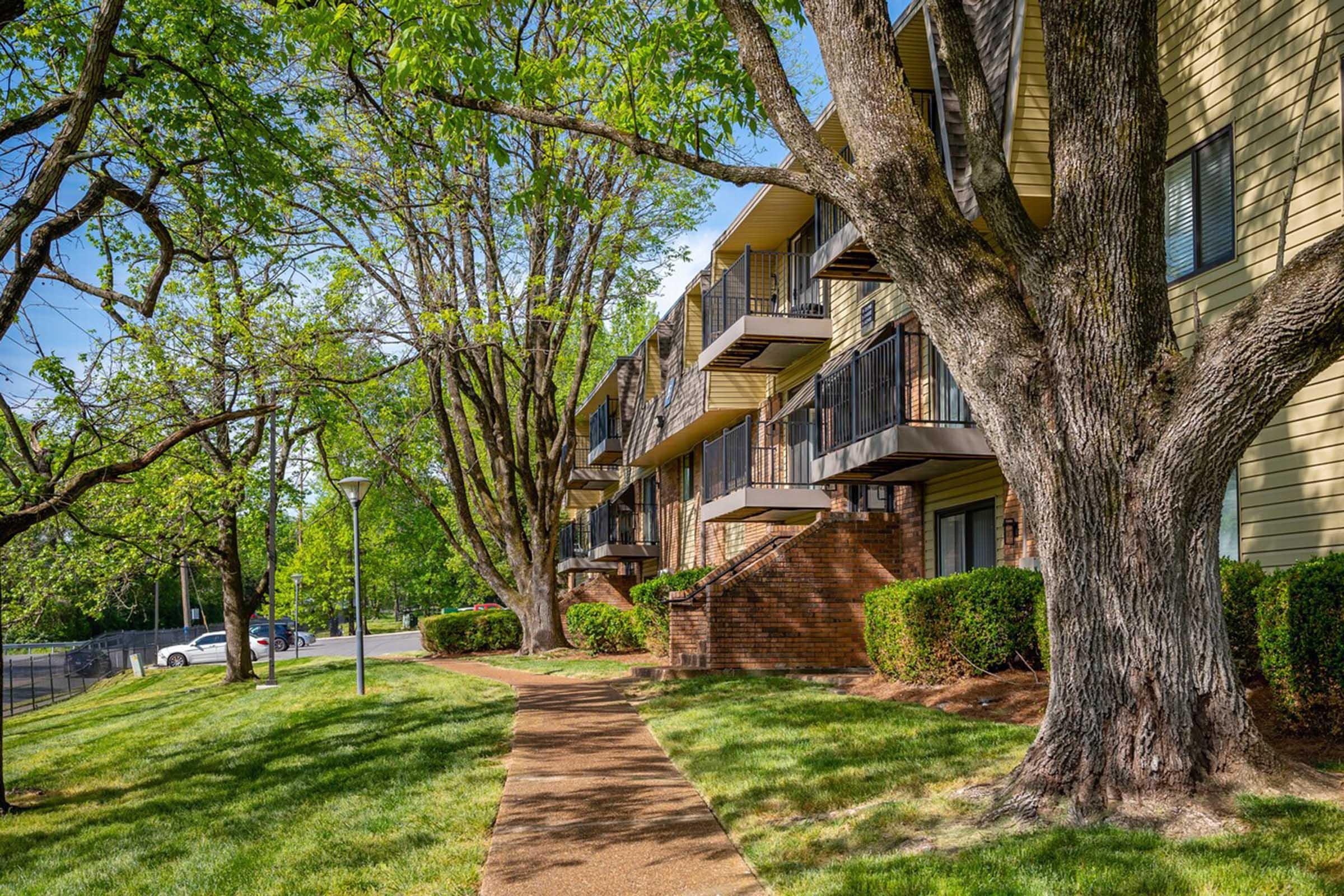 A sunny scene of an apartment building surrounded by lush trees and well-maintained landscaping. A pathway leads up to the building's entrance, with balconies visible on the upper floor. The greenery and clear blue sky create a welcoming atmosphere.