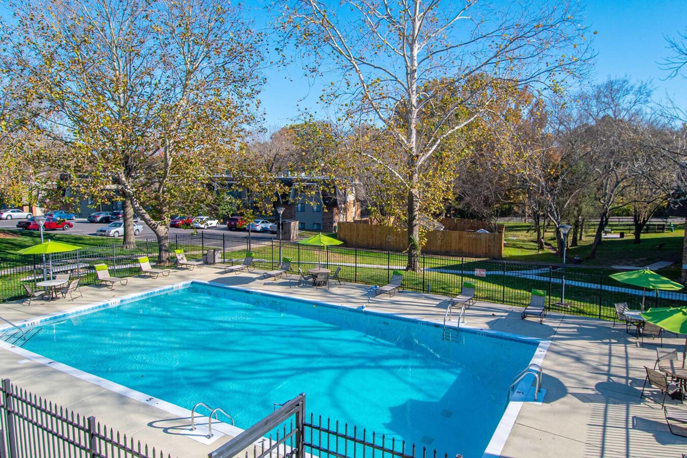 A spacious outdoor swimming pool surrounded by lounge chairs and shaded by umbrellas, set in a landscaped area with trees and a grassy lawn. Clear blue skies above enhance the serene atmosphere.