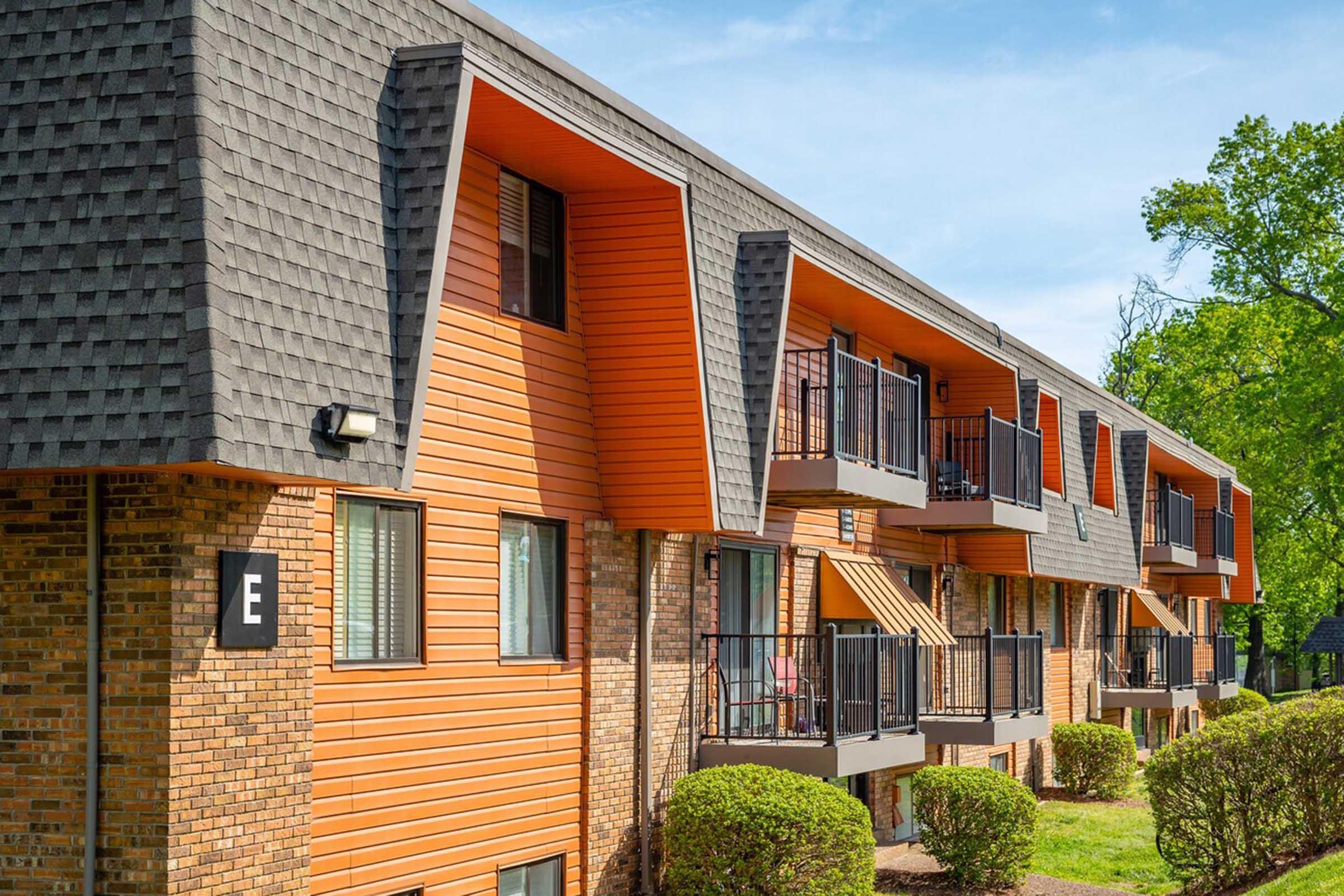 Exterior view of an apartment building featuring a modern design with orange paneling and sloped roofs. The building has multiple balconies with black railings, lush green landscaping in front, and clear blue skies in the background. The scene conveys a welcoming residential atmosphere.