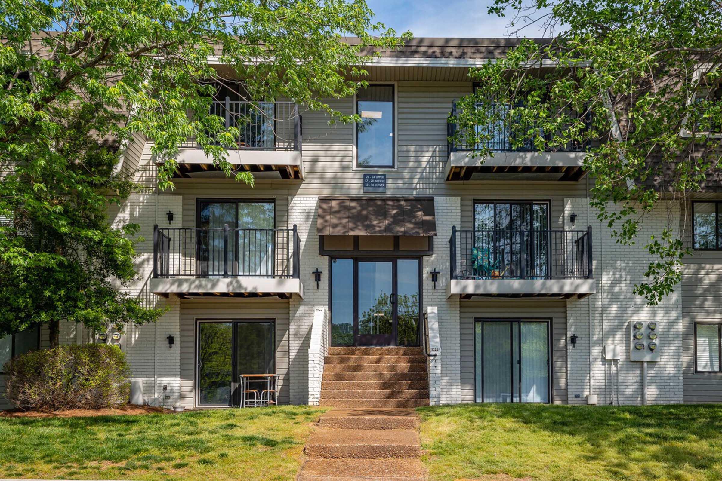 A two-story apartment building with a brick exterior. The entrance is marked by a double door, flanked by large windows and balconies on the upper floors. Green trees and grass surround the entrance, and there are small outdoor seating areas visible.