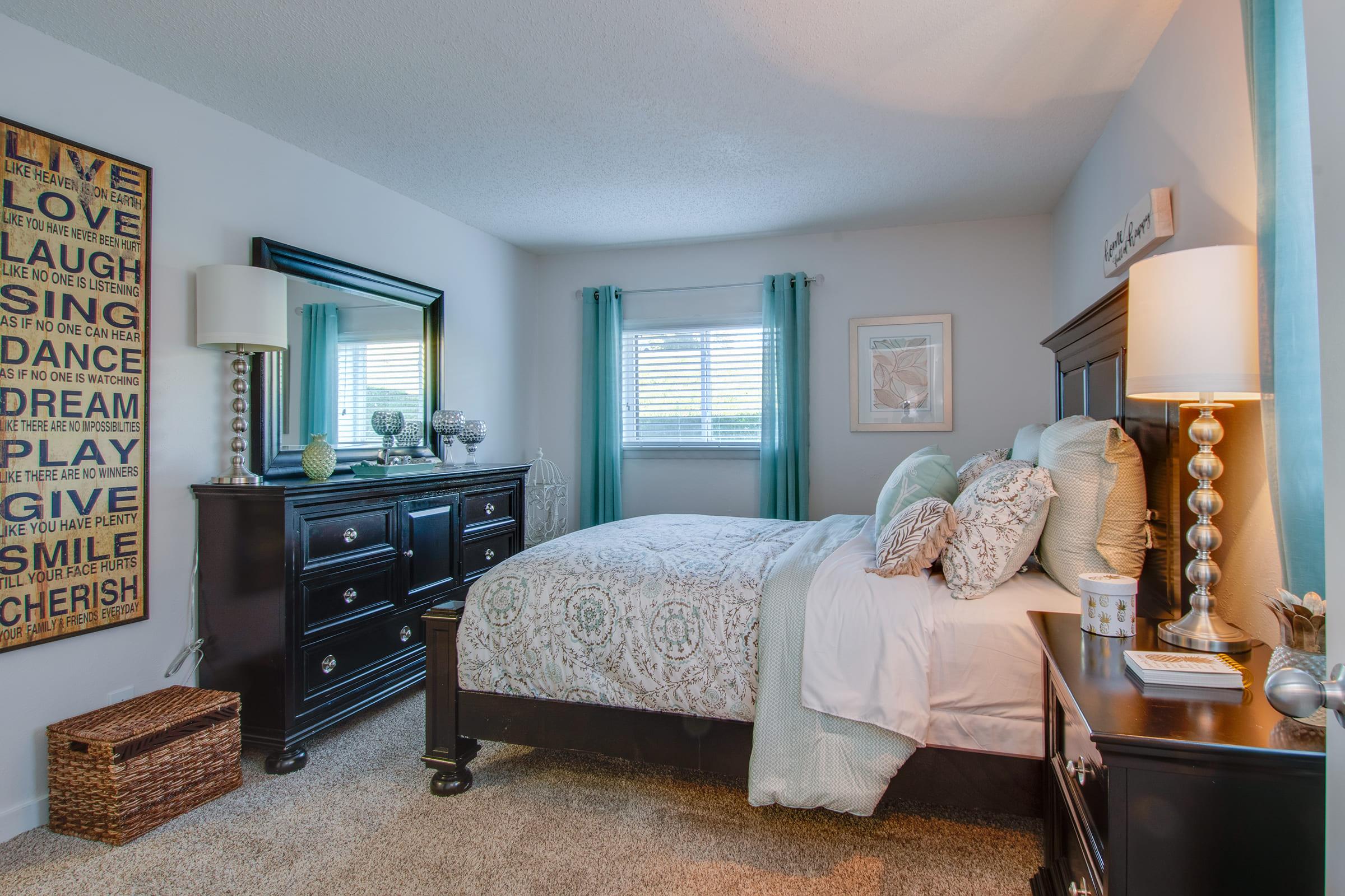 A cozy bedroom featuring a queen-sized bed with light blue and white bedding, a dark wooden dresser with multiple drawers, a large mirror, and decorative items. Natural light flows in through a window dressed with curtains, and the walls are adorned with framed art and a decorative sign. A woven basket adds a touch of warmth.