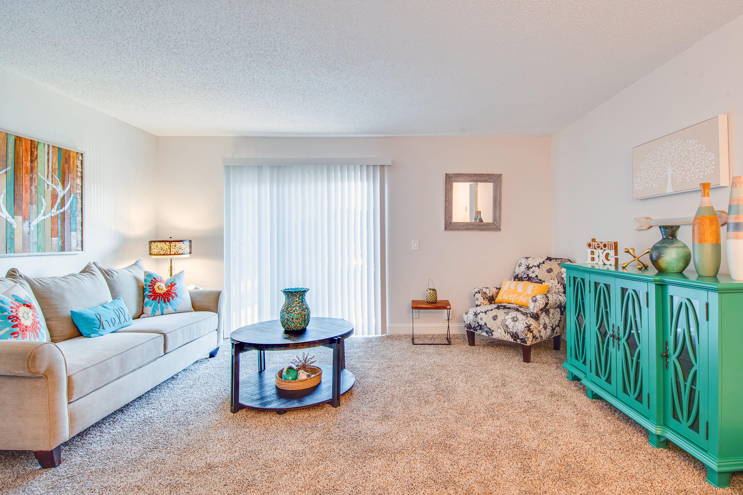 A cozy living room with a light beige sofa adorned with colorful throw pillows. There's a round coffee table in front, topped with a decorative bowl. A teal sideboard holds various decorative items, while a patterned armchair is off to the side. Large windows with sheer curtains allow natural light to brighten the space.