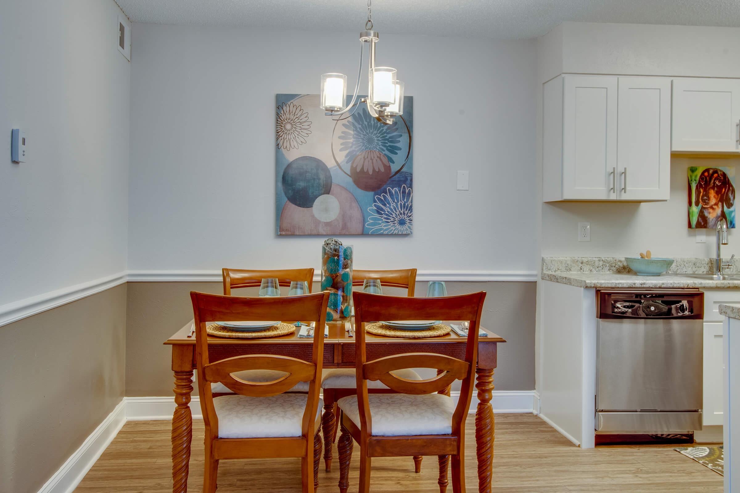 A dining area featuring a wooden table set with plates and glasses, surrounded by four chairs. A decorative painting with abstract floral designs hangs on the wall. A chandelier provides light, and a kitchen area is visible in the background with modern cabinetry and appliances.