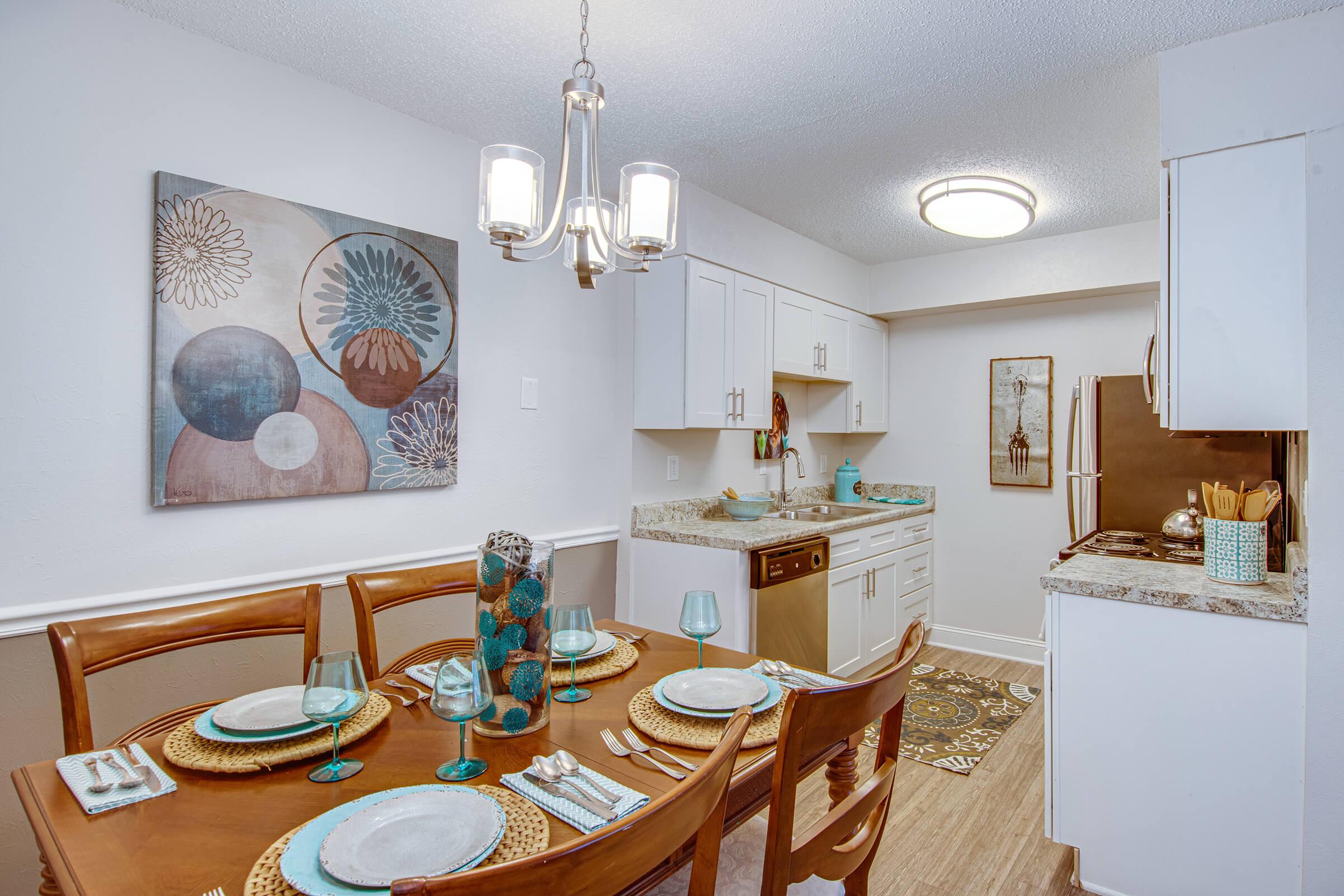 A modern kitchen and dining area featuring a wooden table set for six, with blue glassware and decorative placemats. The kitchen includes white cabinets, a stove, and a granite countertop. Wall art and a floral centerpiece add a stylish touch to the space, illuminated by a contemporary chandelier.