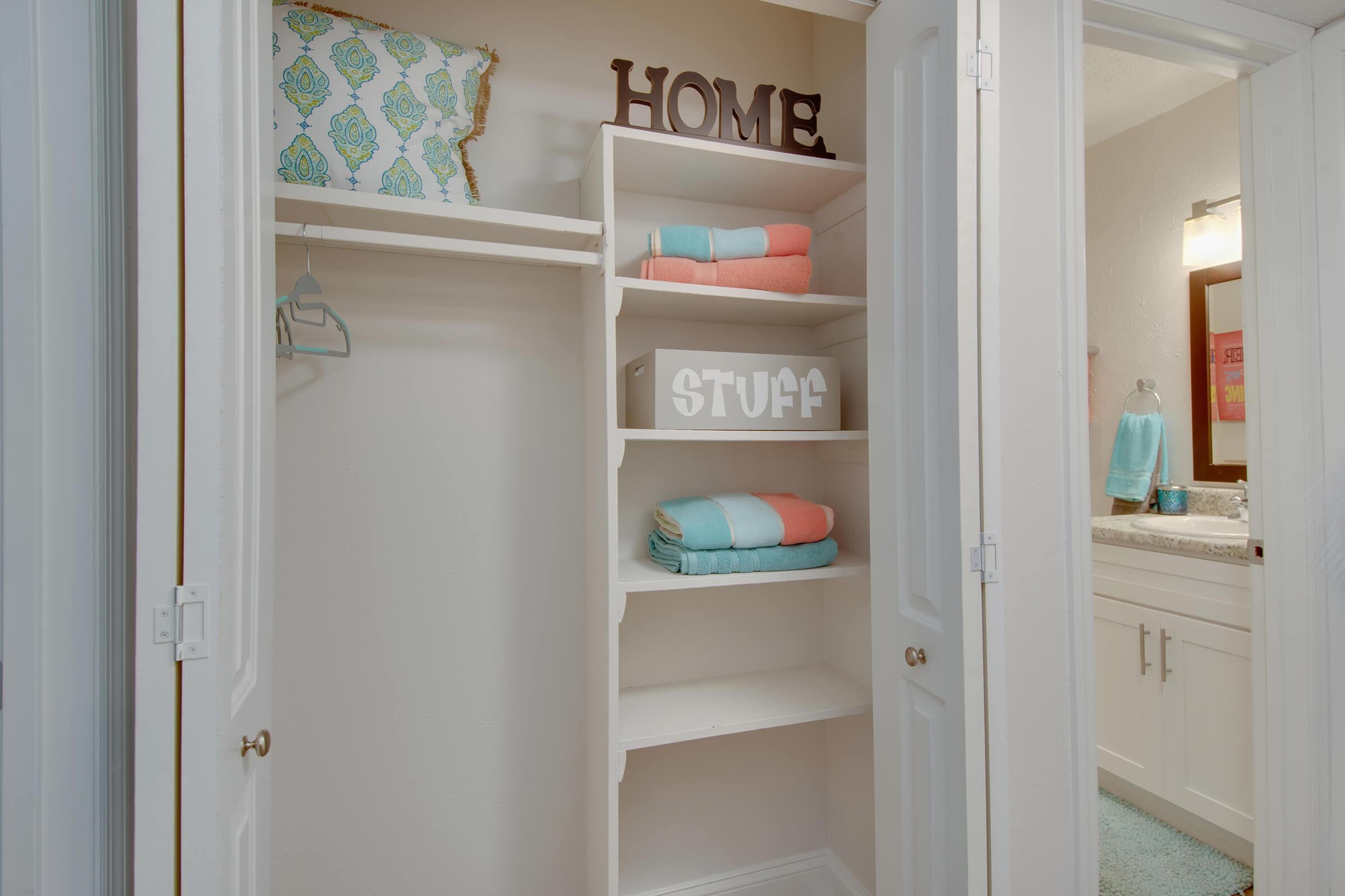 A neatly organized closet with white shelves. On the top shelf, there is a decorative pillow with a blue and green pattern. The middle shelves hold neatly folded towels in coral and aqua colors, and a storage box with the word "STUFF" on it. The closet features double doors and is well-lit.