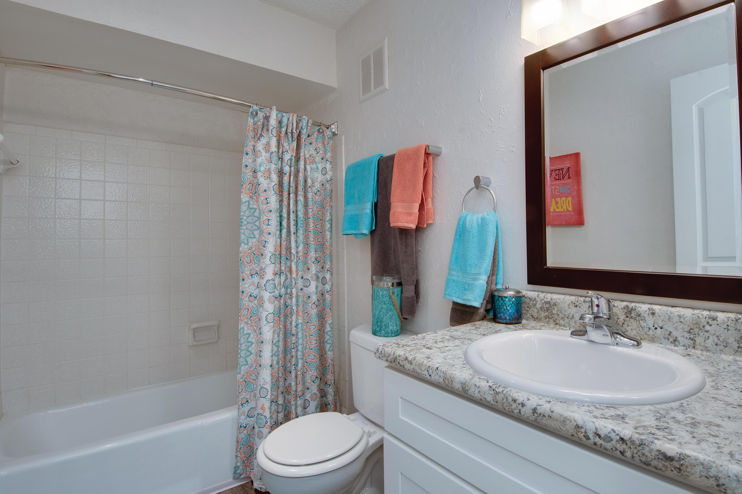 A bright and modern bathroom featuring a bathtub with a shower curtain, a white vanity with a granite countertop, a round sink, and a mirror. Brightly colored towels hang on towel bars and hooks, adding vibrant accents to the neutral walls and decor.
