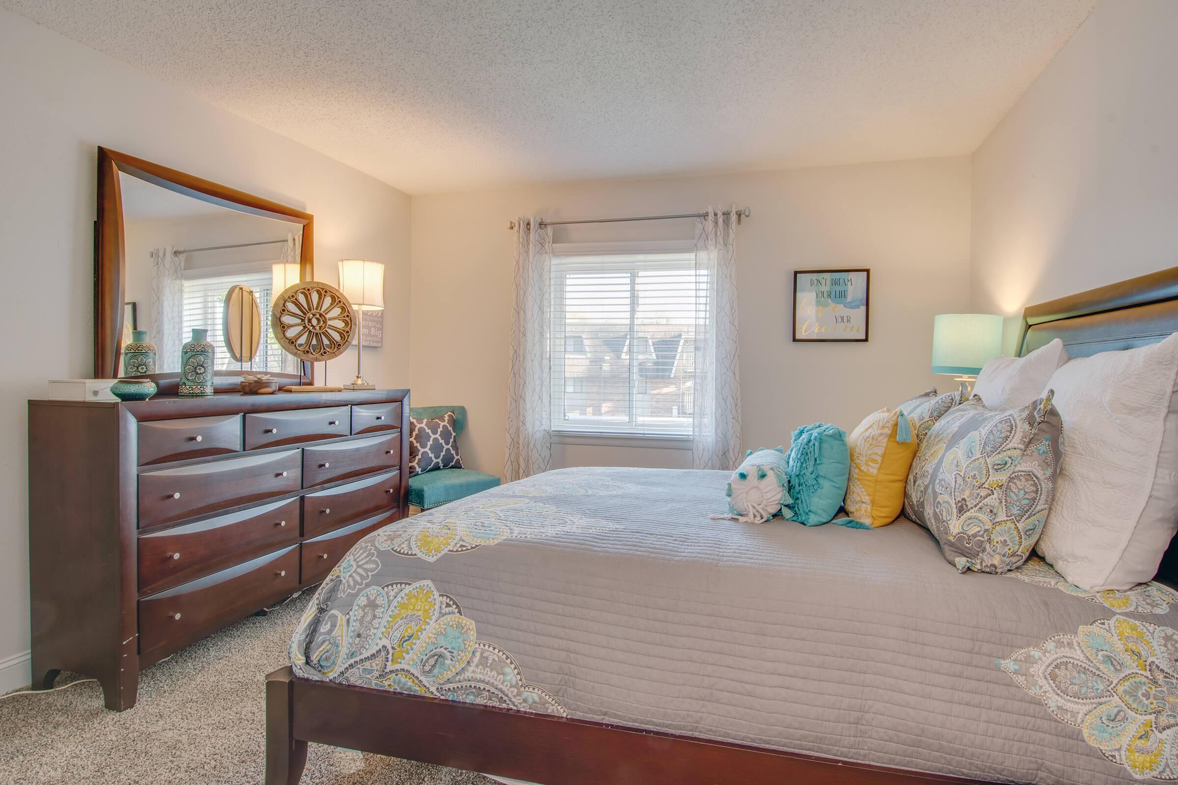 A cozy bedroom featuring a bed with decorative pillows and a gray quilt. There's a dark wooden dresser with decorative items, a large mirror above it, and two lamps providing soft light. A window offers natural light, and a small blue armchair is positioned near the window. The décor includes shades of blue and yellow.