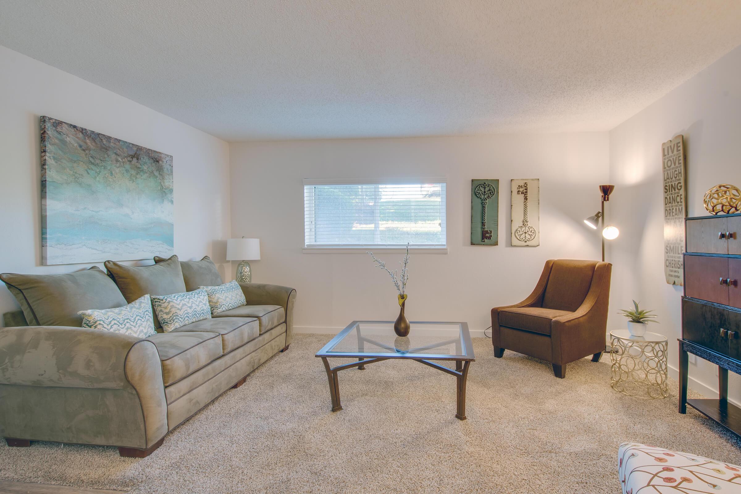 A cozy living room featuring a green sofa adorned with decorative pillows, a glass coffee table, and an armchair. A small decorative plant and a cabinet are nearby, with wall art and a lamp adding character. Natural light streams through a window, creating a warm and inviting atmosphere.