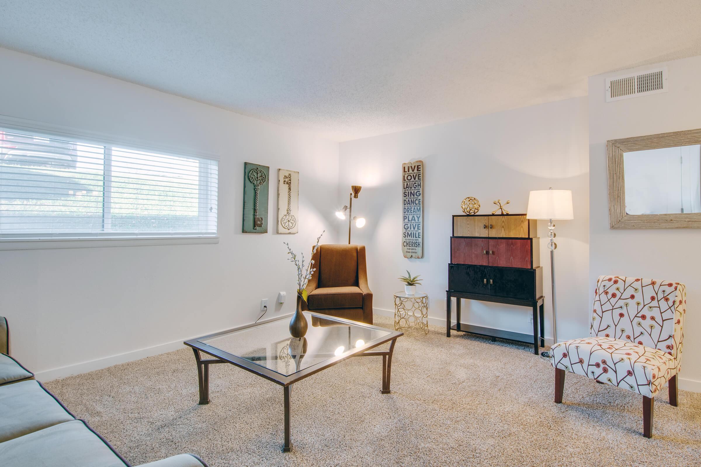 A cozy living room featuring a beige sofa, a glass coffee table, and two armchairs. One armchair is upholstered in a dark fabric, while the other has a floral pattern. A lamp stands beside the sofa, and a short cabinet with colorful drawers is against the wall. Decorative items and plants add a cozy touch.