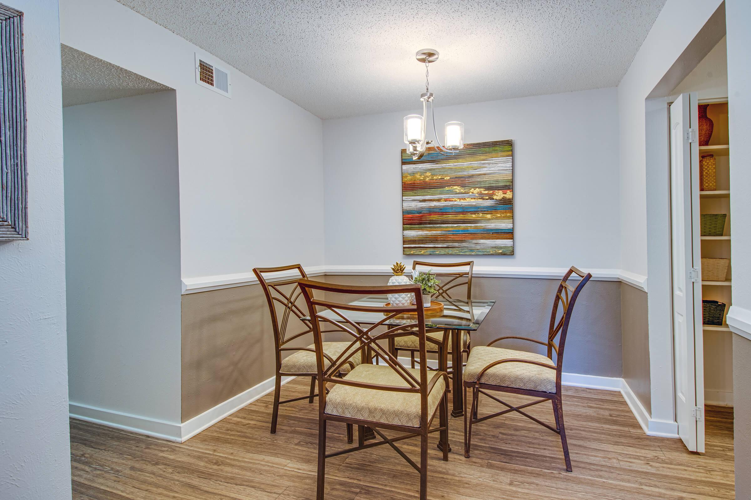 A bright dining area featuring a glass-top table surrounded by four upholstered chairs. The walls are painted in light colors, and there is a colorful abstract painting above the table. A stylish light fixture hangs from the ceiling, and there are built-in shelves visible in a nearby nook.