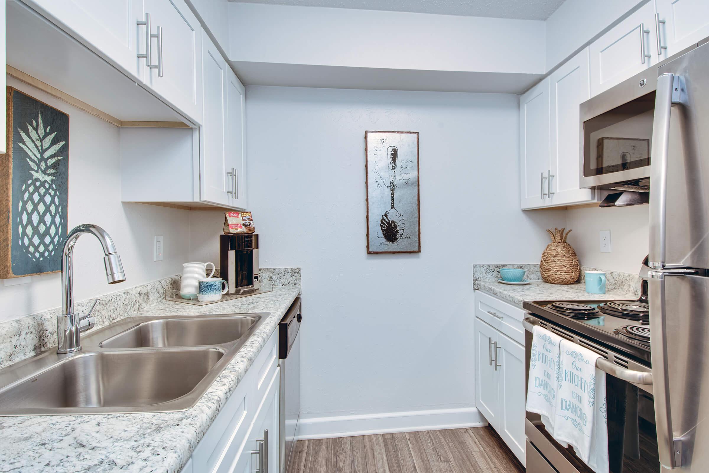 Modern kitchen featuring white cabinetry, a granite countertop, and stainless steel appliances. There are two sinks, a coffee maker, and decorative elements including a pineapple artwork and a metal wall hanging. Light-colored walls and wood-style flooring complete the inviting atmosphere.