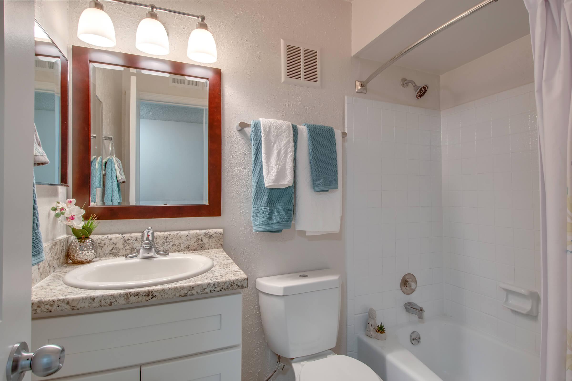 A clean and modern bathroom featuring a white bathtub with a shower, a light-colored vanity with a granite countertop, a mirror with a wooden frame, and neatly arranged towels in turquoise and white. A small potted plant and a decorative flower add a touch of nature.