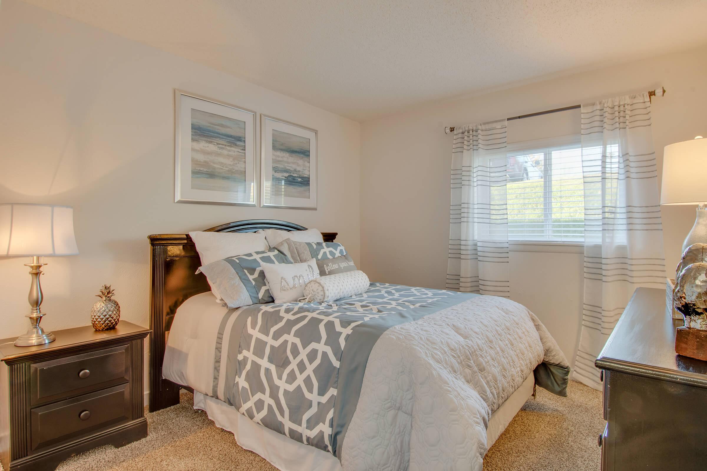 A cozy bedroom featuring a queen-sized bed with gray and white bedding, decorative pillows, and two framed prints on the wall. There's a nightstand with a lamp and a small pineapple decoration, as well as a dresser beside the window, which is covered with light, sheer curtains allowing natural light to enter.