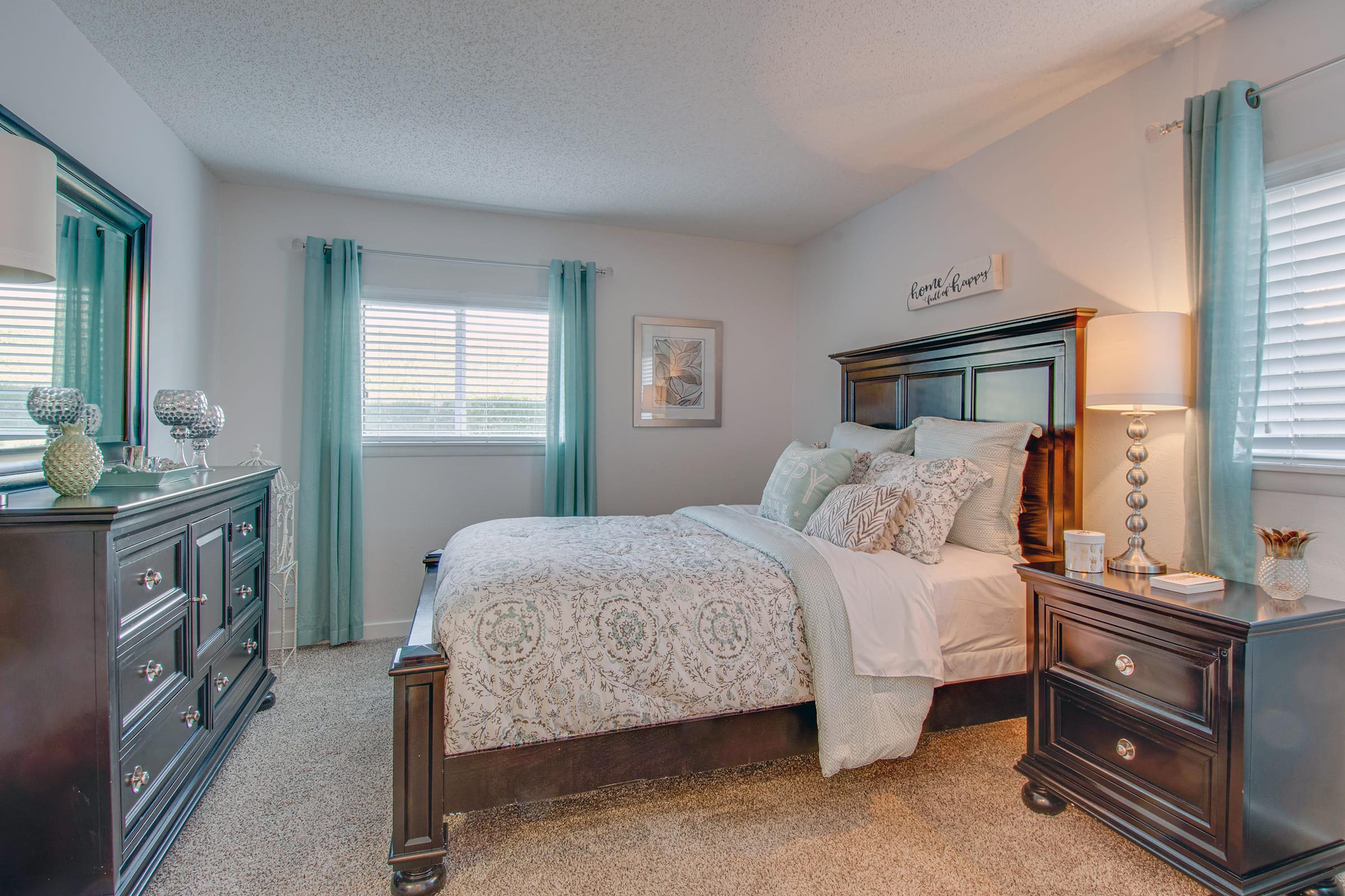 A cozy bedroom featuring a bed with patterned bedding, decorative pillows, and a plush throw. A dark wood dresser and nightstand are present, along with a lamp and decorative items. Soft blue curtains frame the windows, allowing natural light to enhance the warm atmosphere.
