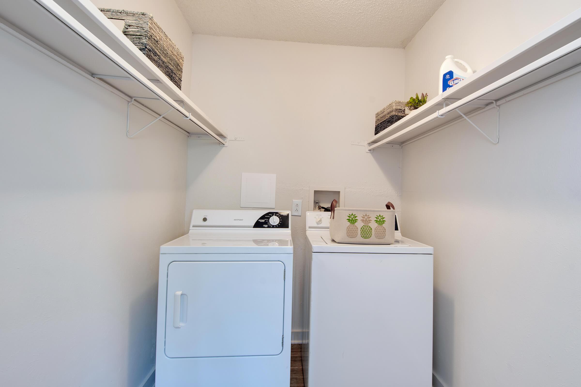A clean laundry room featuring a stacked washer and dryer, with open shelving above. The shelves hold neatly stacked items, while a decorative container with a pineapple design sits on the counter next to the appliances. The walls are plain and the flooring is a light wood.