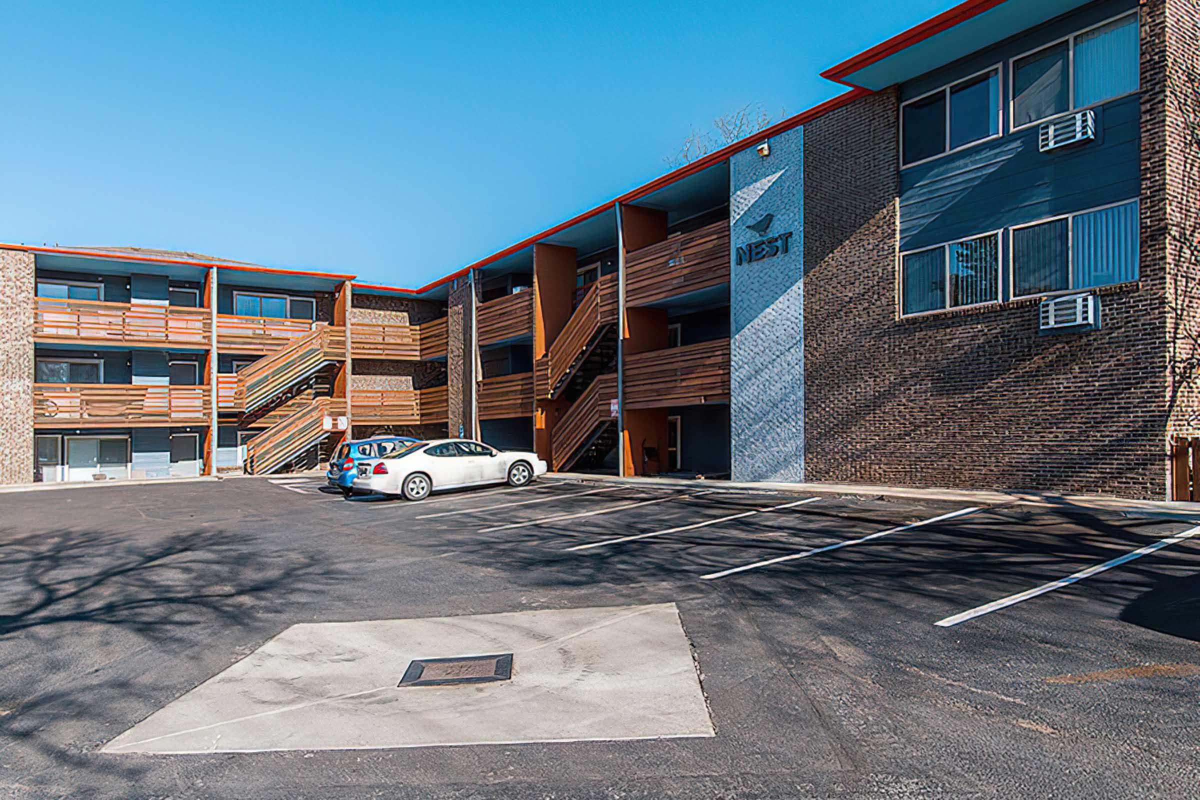 A view of a three-story apartment building with brick and wooden exterior. The entrance features multiple staircases. In the foreground, there is a paved parking lot with parked cars. The building has air conditioning units visible in several windows, and the name "NEST" is displayed on the wall. Clear blue sky above.