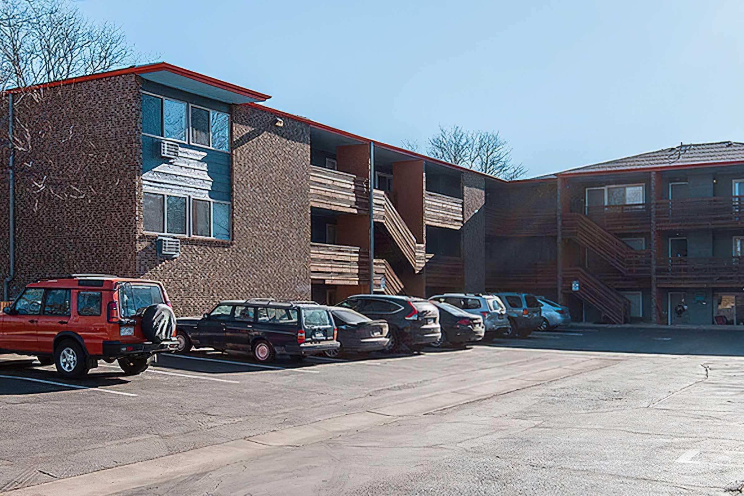 A multi-story brick apartment building with red accents is shown. There are several parked cars in the foreground, including a red SUV and a dark sedan. The scene is set in a sunny outdoor environment with a blue sky. The building features balconies and air conditioning units on the exterior.